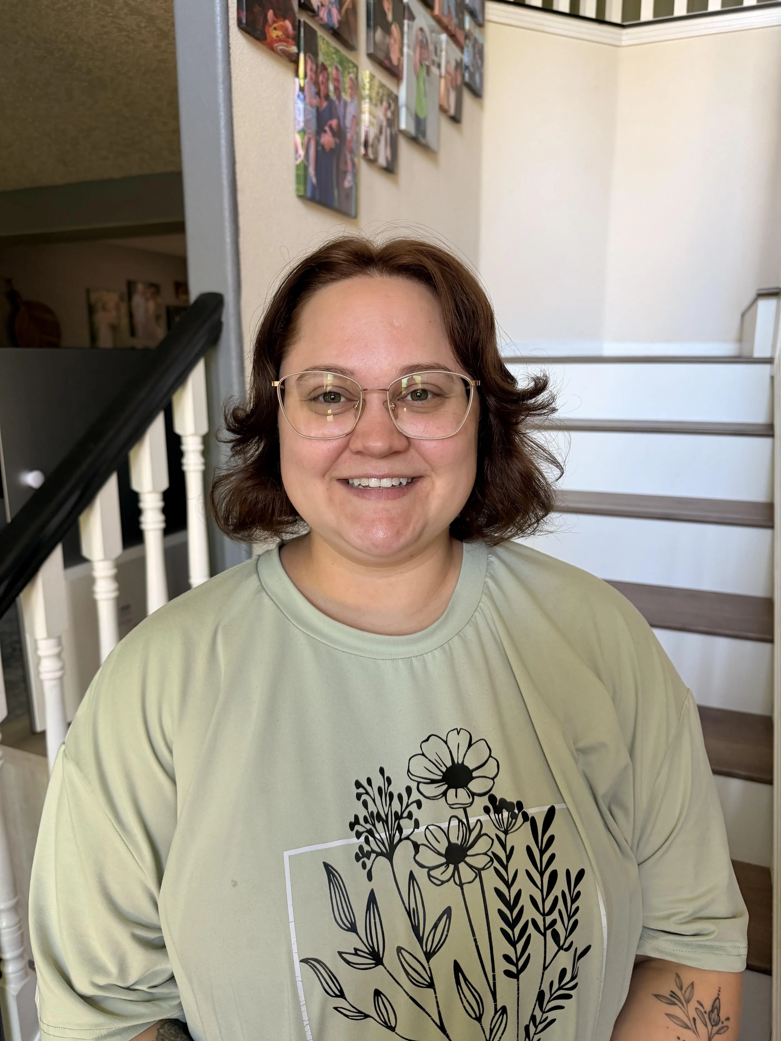 Picture of Isabella who is has shoulder length brown hair, is wearing glasses, and a sage green tee with graphic flowers in black.