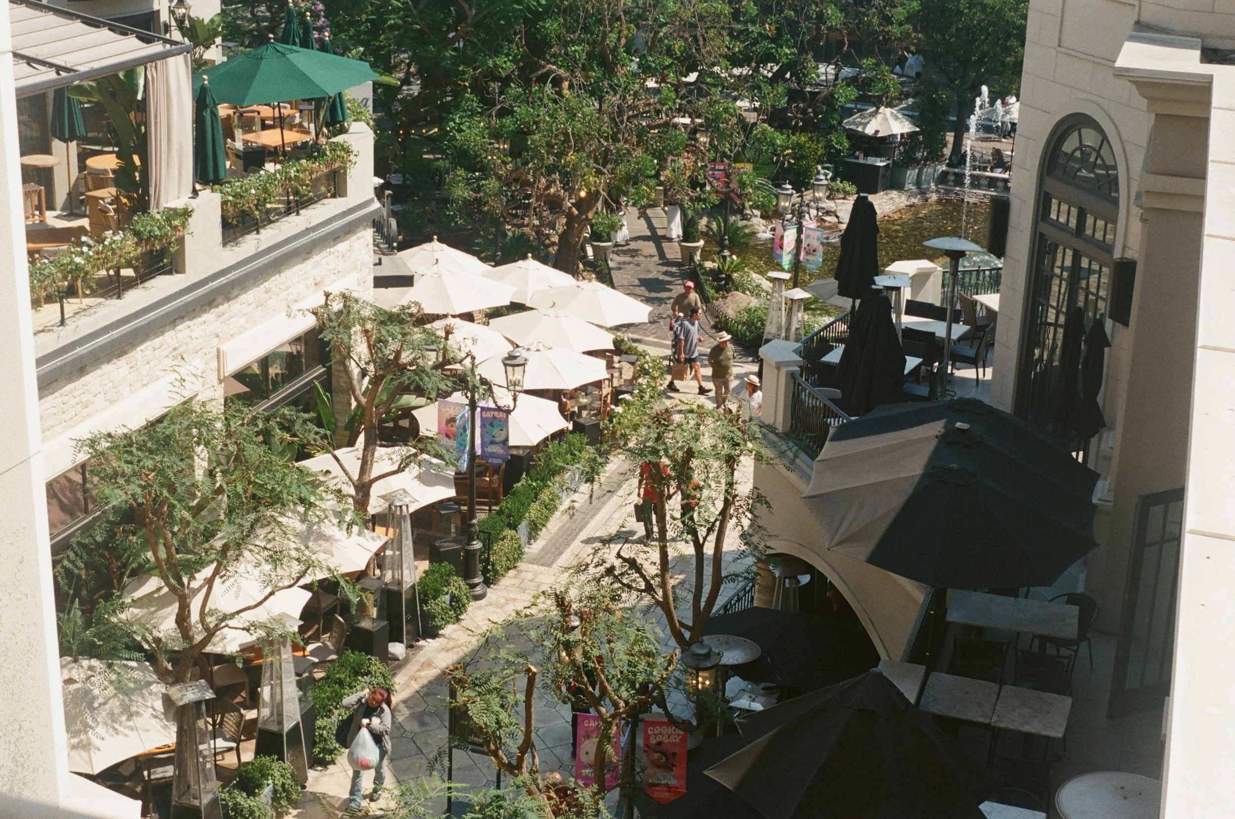 Outdoor shopping and dining area with parasols, people walking, and trees.