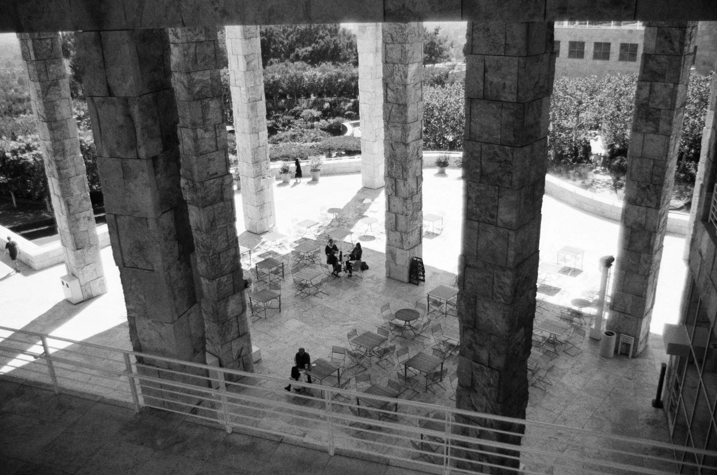 Black and white photo of an outdoor patio area with several tables and chairs, large stone columns, and a few people sitting and walking around.