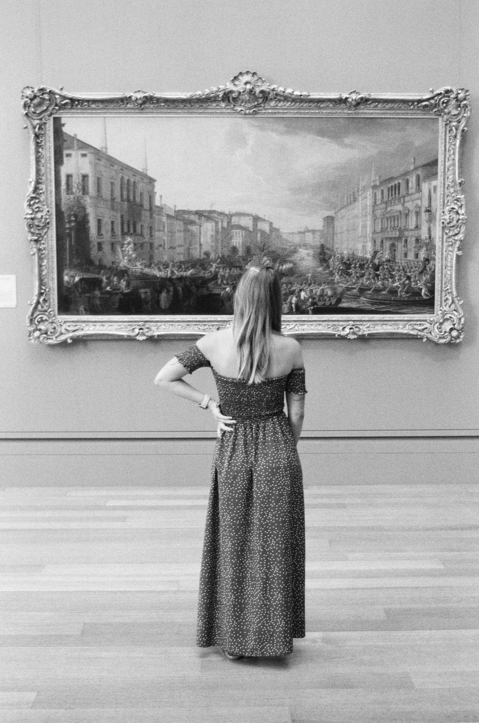 A woman with long hair in an off-the-shoulder dress standing in an art gallery, looking at a large framed painting of a city street scene.