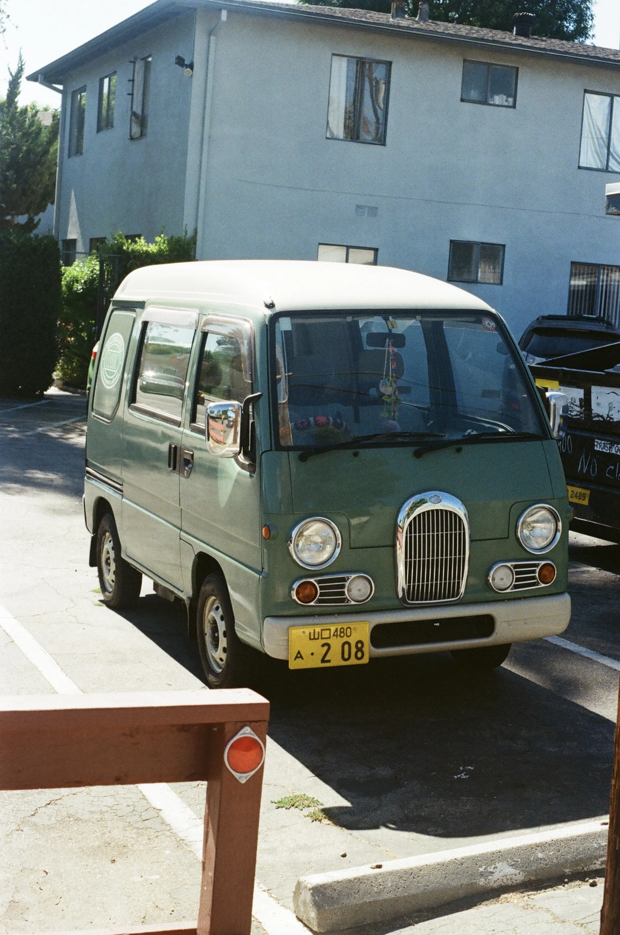 A small green vintage kei van parked in a parking lot near a residential building. The van has a rounded front, chrome details, and a yellow license plate with Arabic characters. Shadows indicate sunny weather.