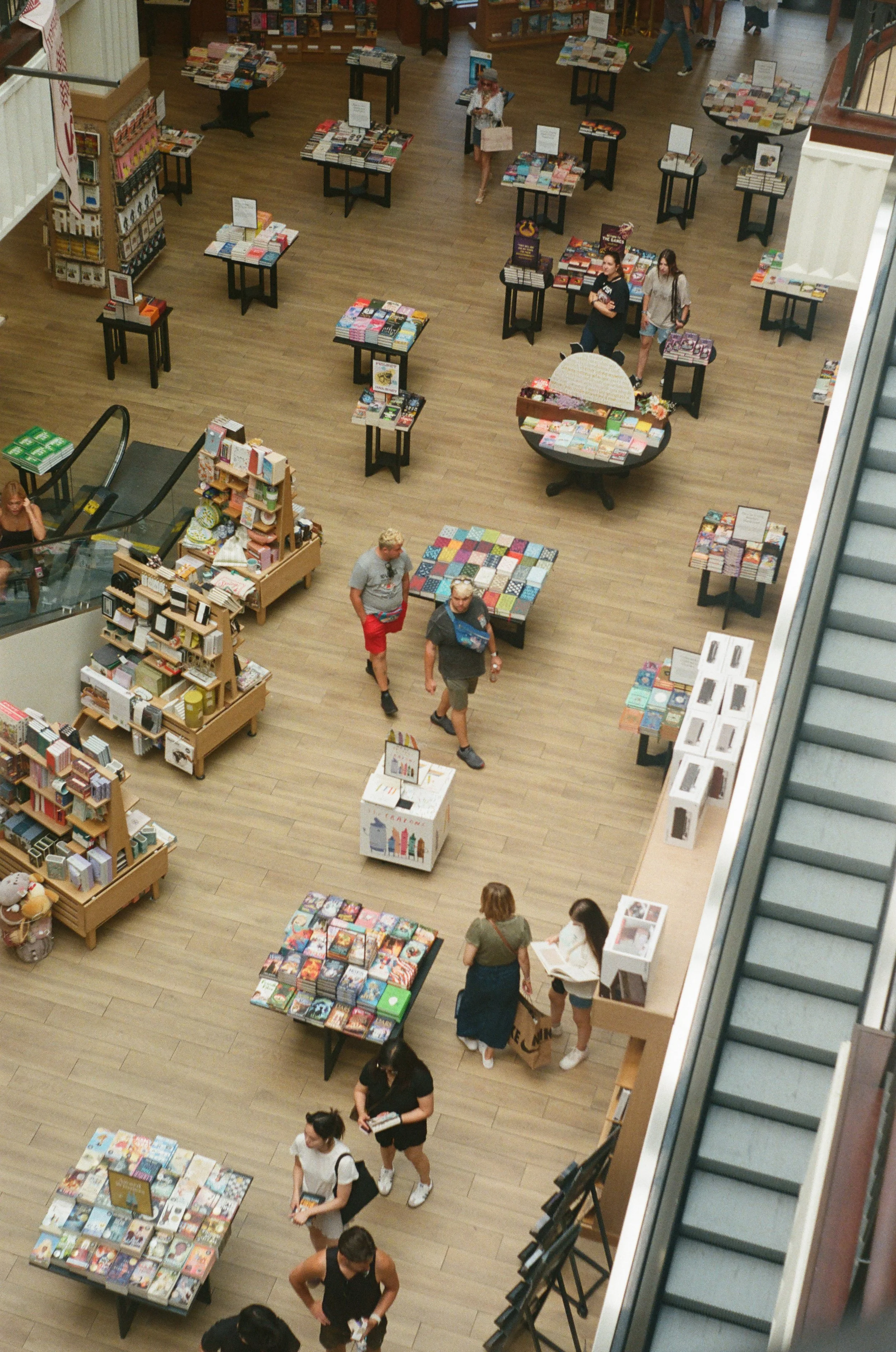 Bird's eye view of a bookstore with multiple tables and shelves displaying books, several shoppers browsing, and an escalator on the right side.