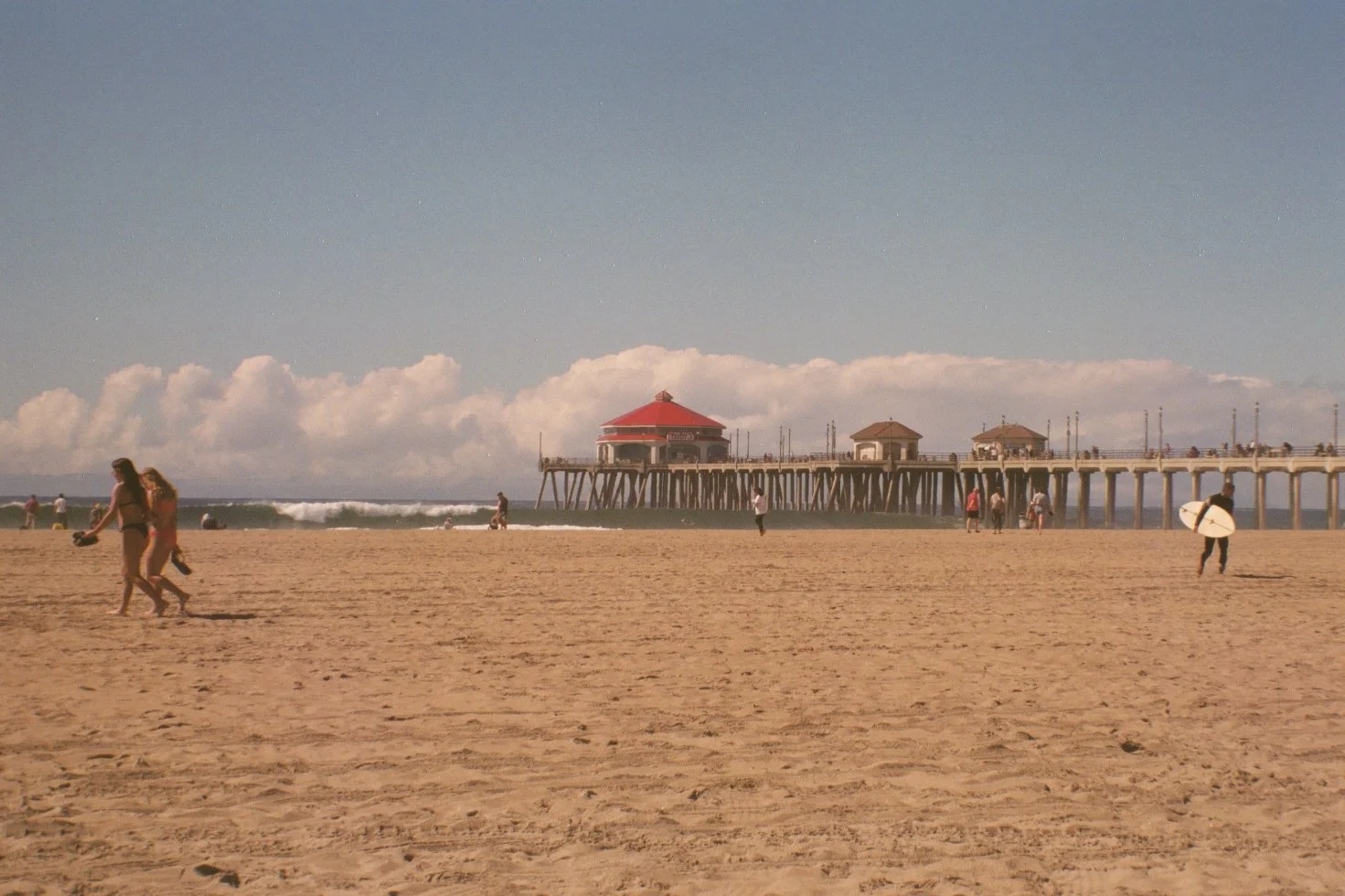 Beach scene with people walking on sand, some holding surfboards, a pier with red and brown structures extending into the ocean, and waves in the background under a partly cloudy sky.
