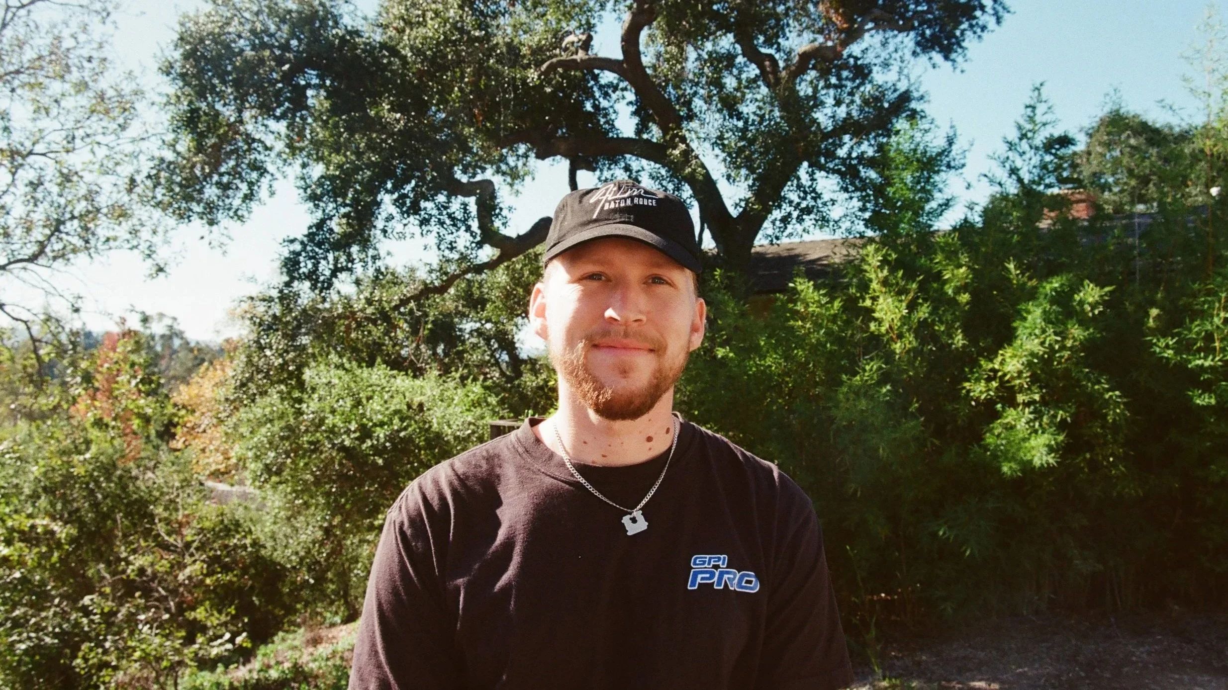 A man with a beard and mustache wearing a black cap and black T-shirt standing outdoors with trees and bushes in the background.
