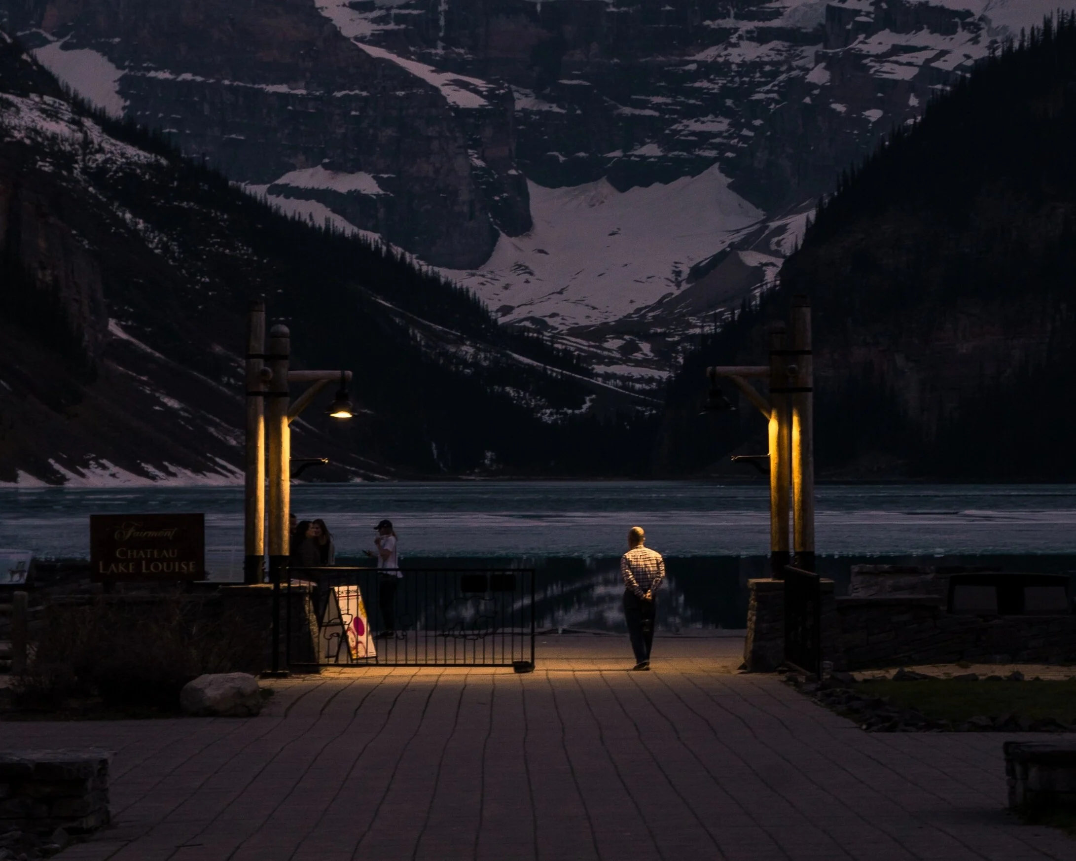 People gathered at scenic lakeside viewing area during dusk, with mountains and snow patches reflected on the lake