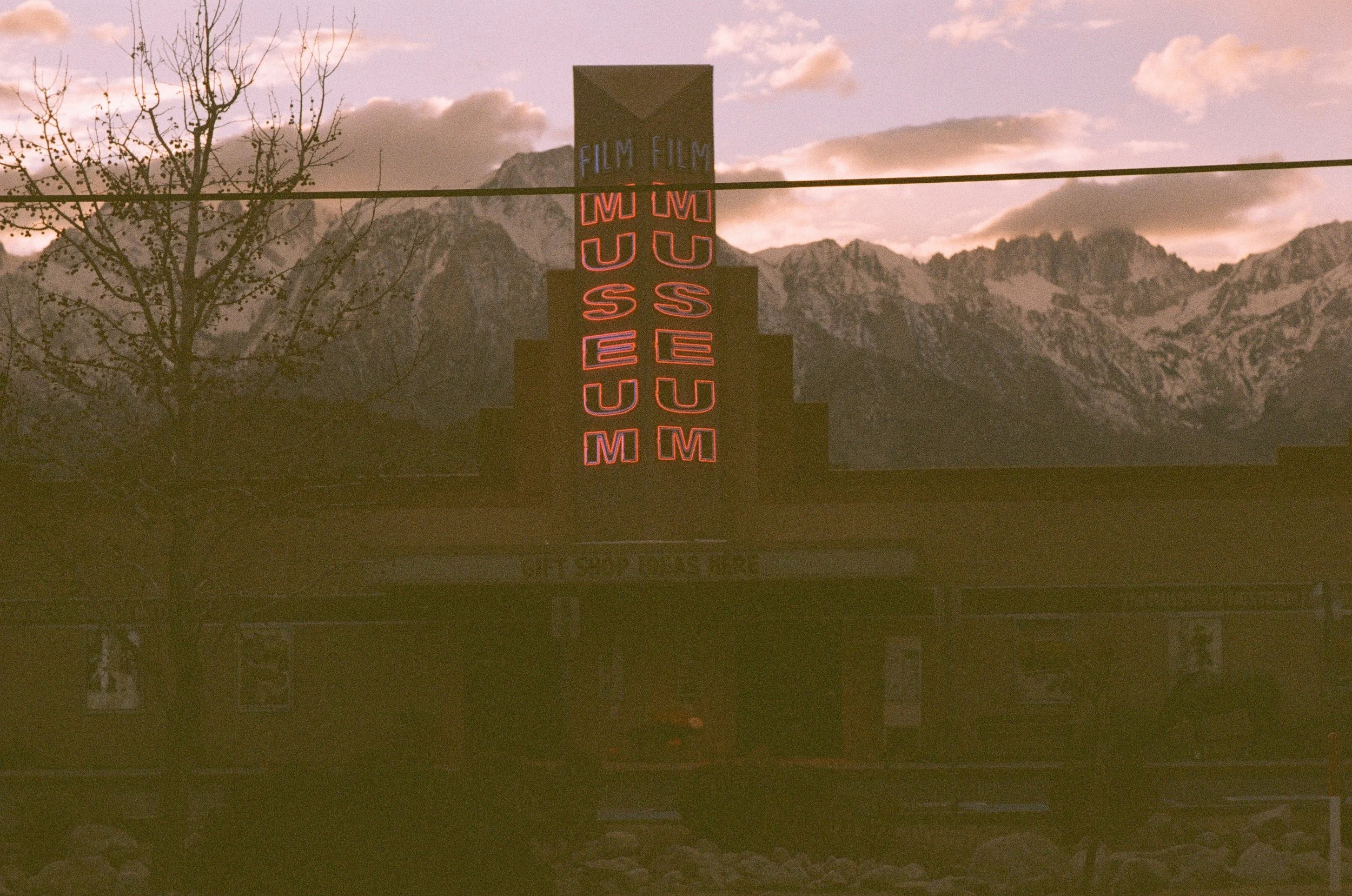A neon sign reading 'MUSEUM' in reverse with the word 'FILM' above it, set against mountains and a cloudy sky at sunset.
