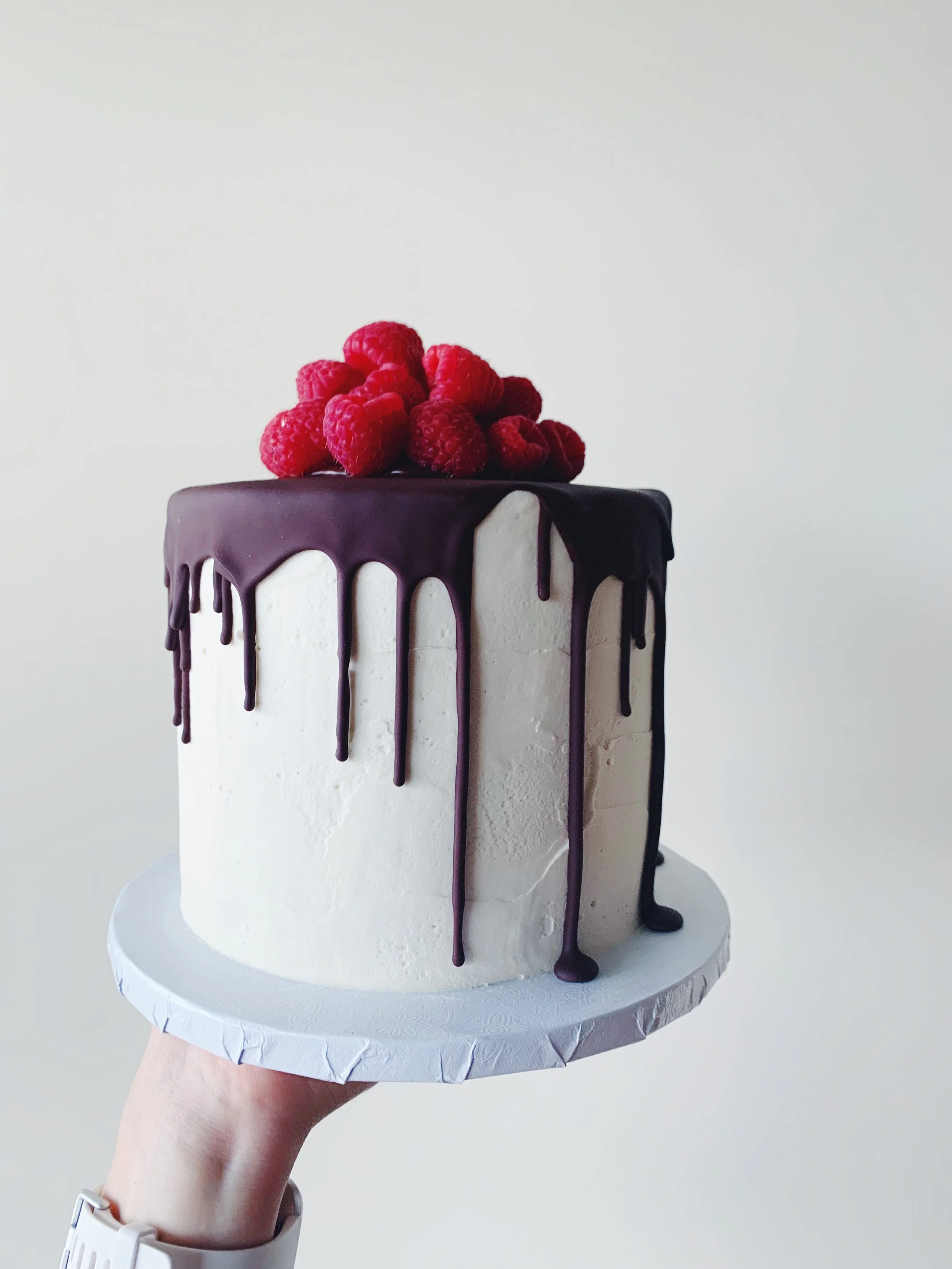 A white frosted cake with dark chocolate ganache drips, topped with fresh raspberries.