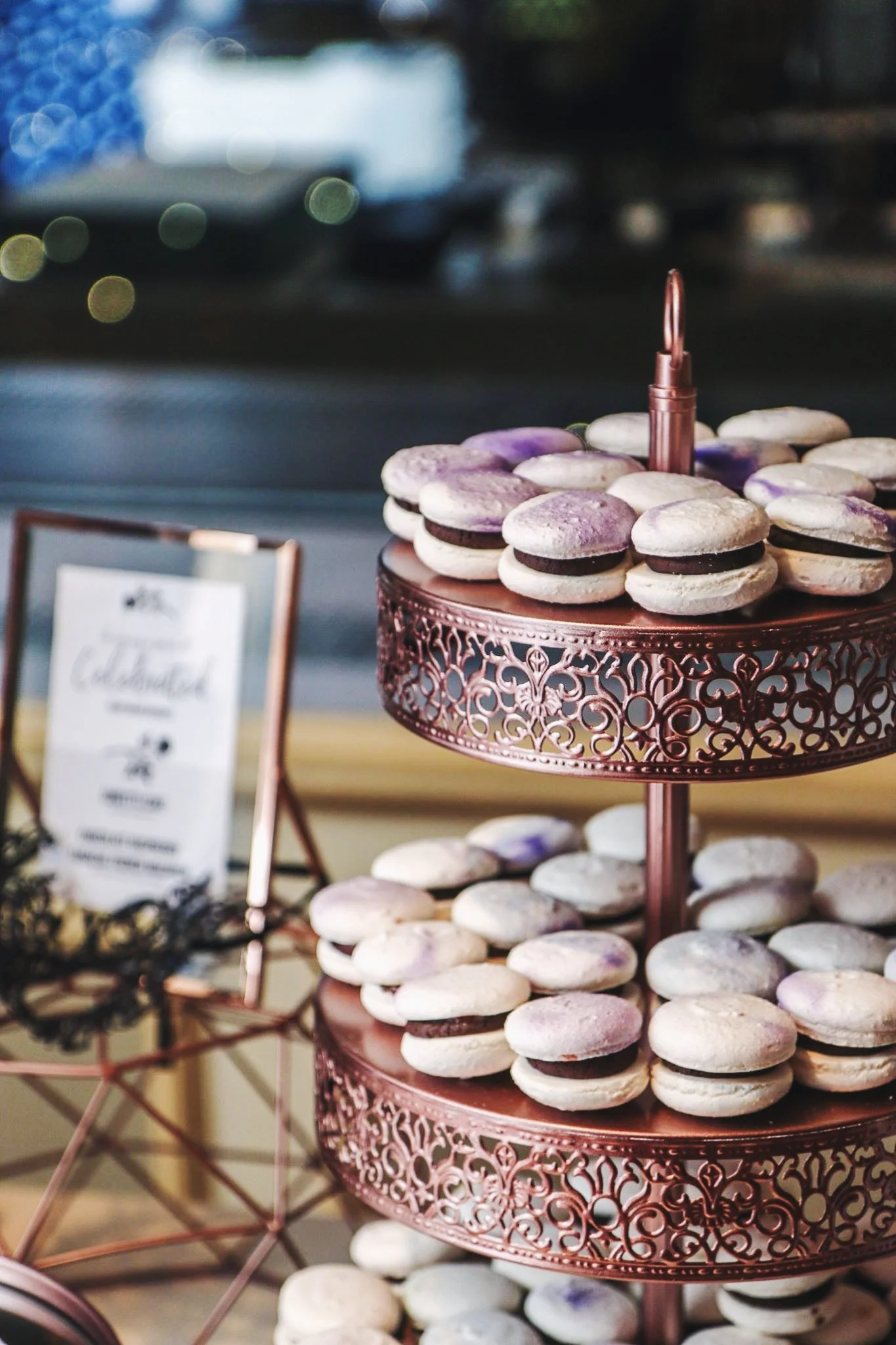 A display of white macarons with purple accents and dark filling on a decorative two-tiered metal stand at an event.
