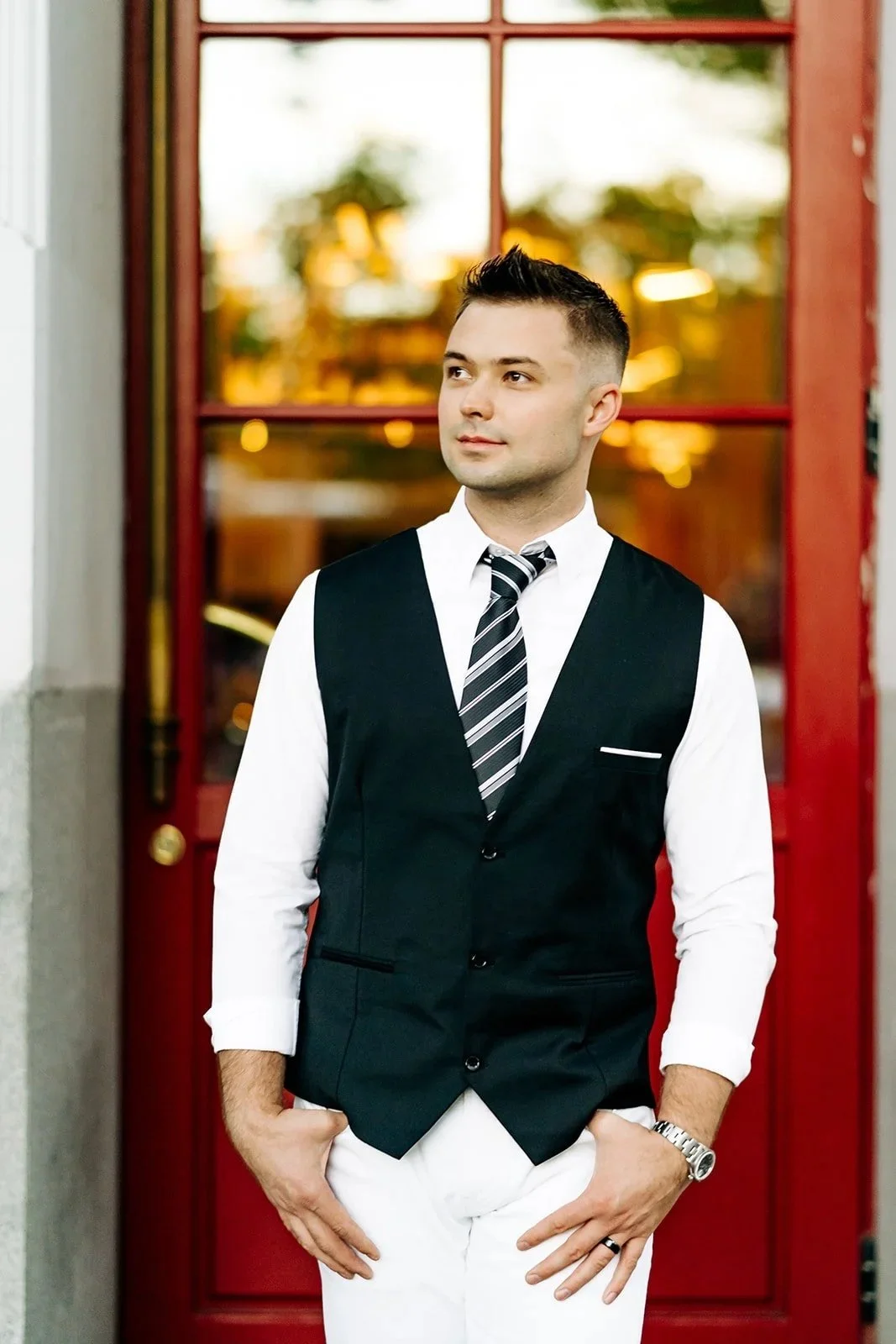 A young man with short dark hair, wearing a white shirt, black vest, striped tie, white pants, and a silver watch, standing in front of a red door with glass panels.