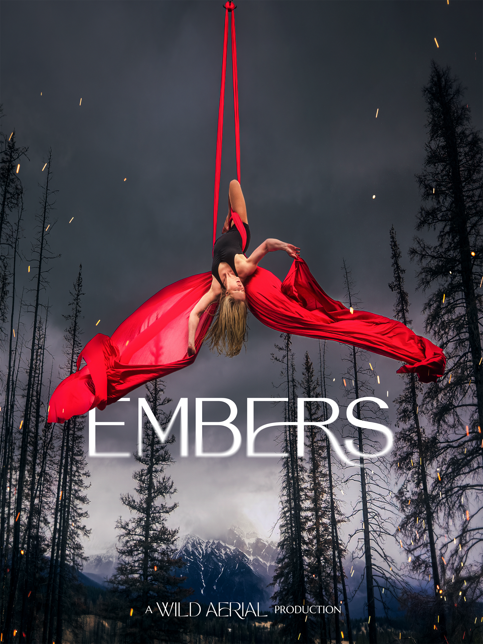 Aerial performer hanging upside down on red silk fabric strung from the sky during a dark mountain landscape with trees and snow-capped peaks, with sparks flying around.