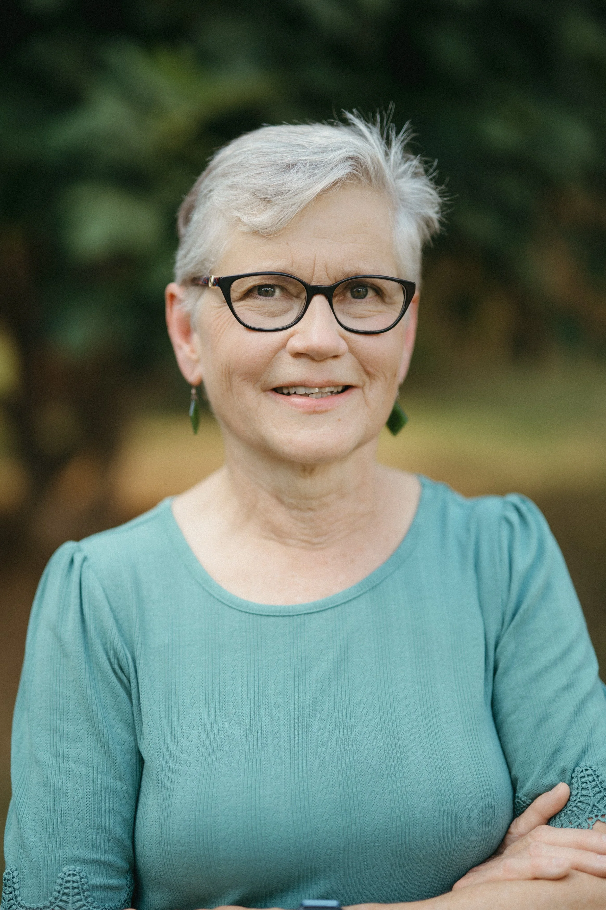 Close-up of an elderly woman with short gray hair, black-framed glasses, green earrings, wearing a teal blouse with embroidered sleeves, standing outdoors with a blurred green background.