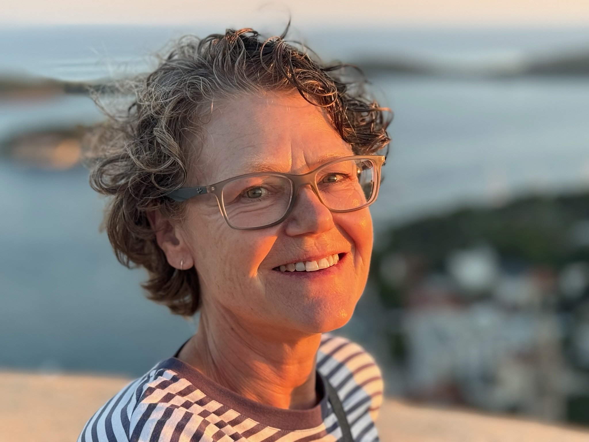 Close-up of a smiling woman with gray curly hair and glasses, wearing a striped shirt, outdoors at sunset near water.