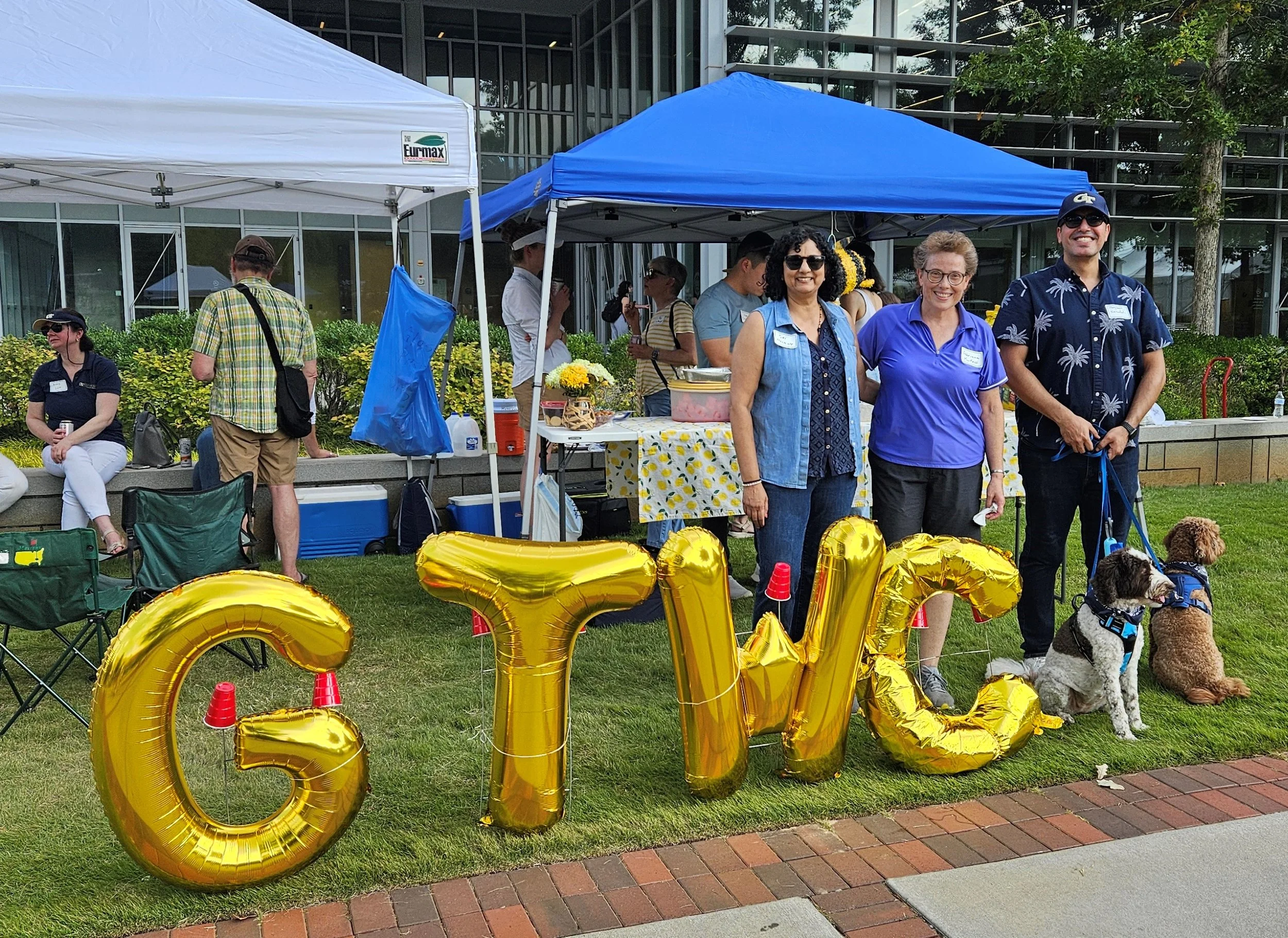 People gathered outdoors at an event with blue and white tents, some sitting and others standing, including three women in the front smiling, and large gold balloons spelling out GTWC.