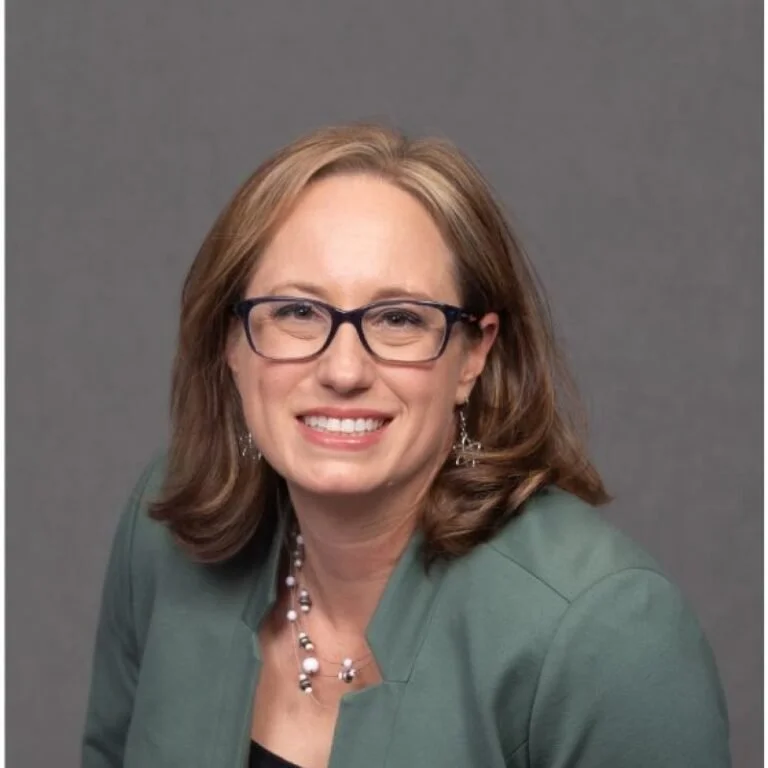 Professional woman with glasses and shoulder-length hair smiling, wearing a green blazer and jewelry, against a plain grey background.