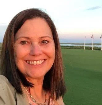 A woman smiling outdoors near a golf course with flags and water in the background.