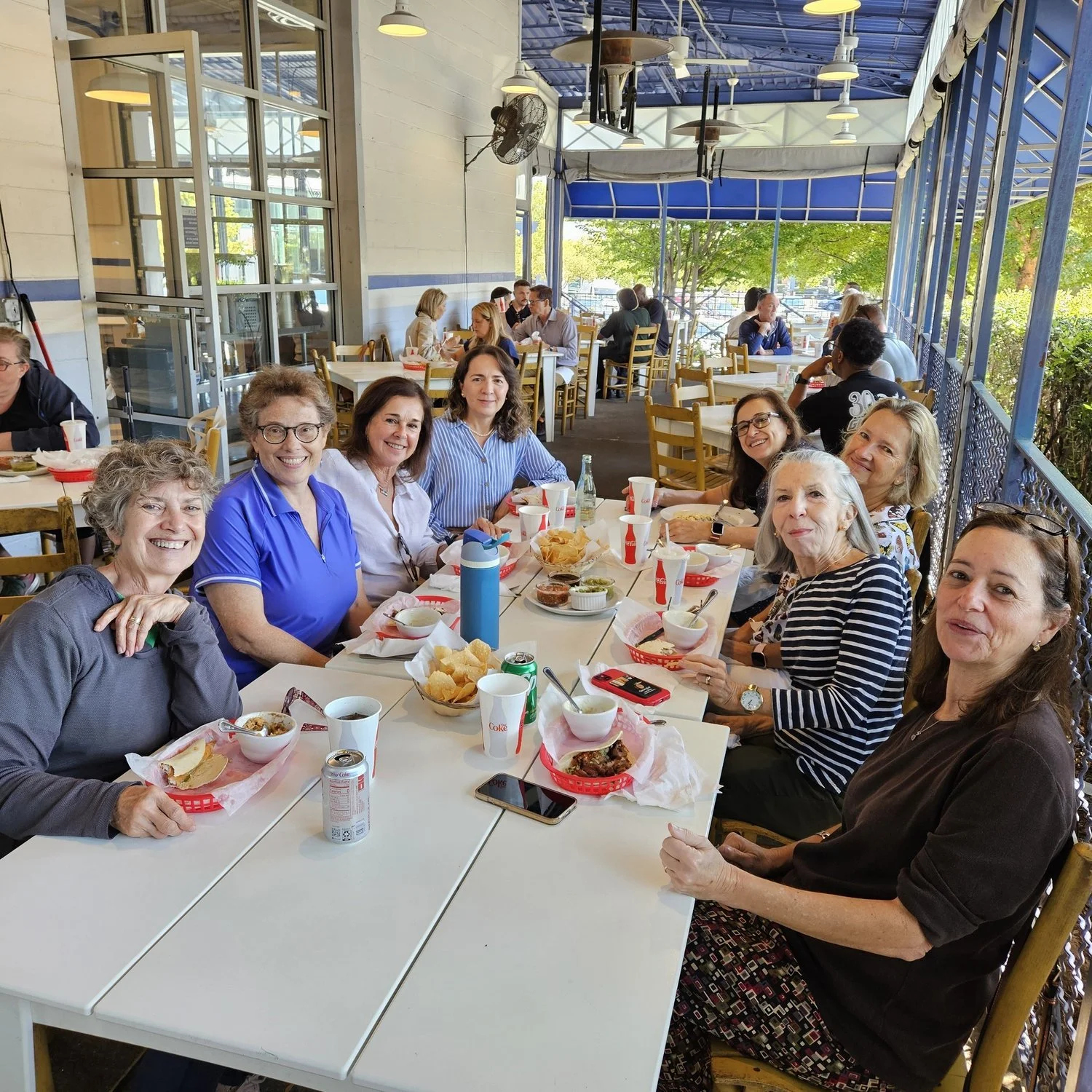 A group of women sitting around a table in a sunny, open-air restaurant, enjoying lunch with chips, salsa, and drinks. This is for a group that meets to speak Spanish.