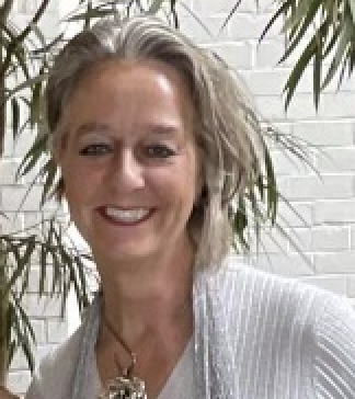 Smiling woman with blonde hair standing in front of a white brick wall and green plants.