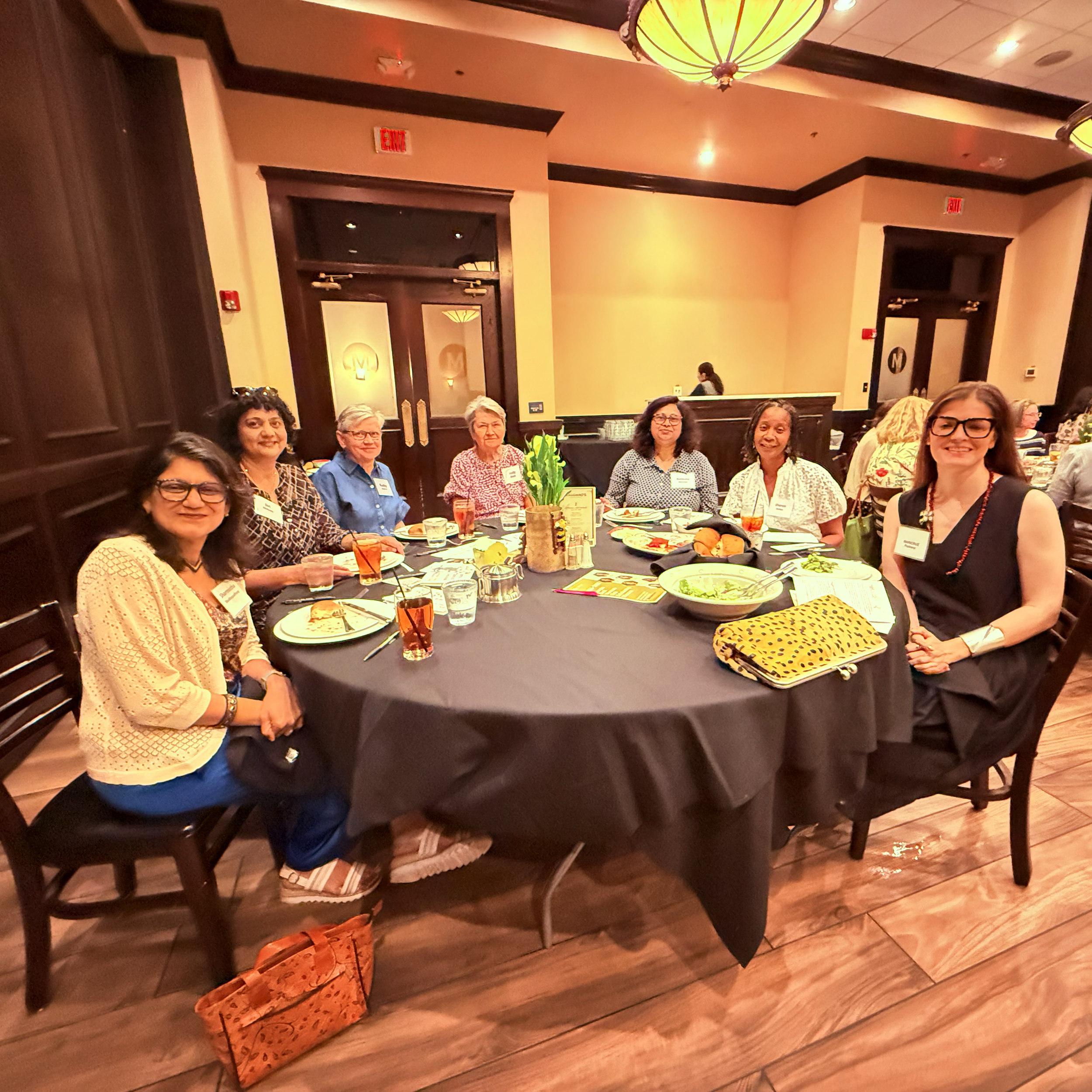 People sitting at a round dining table in a restaurant, smiling for the camera, with plates of food, drinks, and a yellow gift bag on the floor.