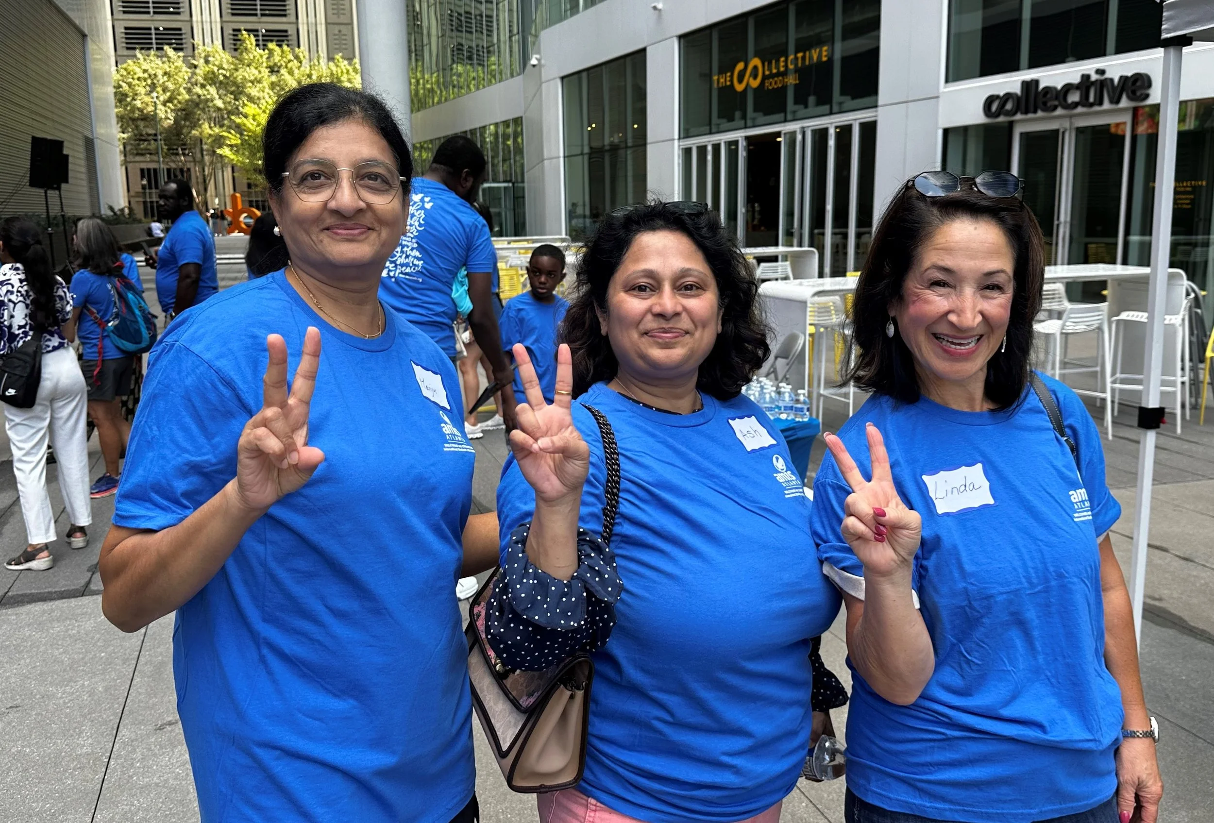 Three women in blue shirts smiling and making peace signs at an outdoor event, with a glass building and people in the background.