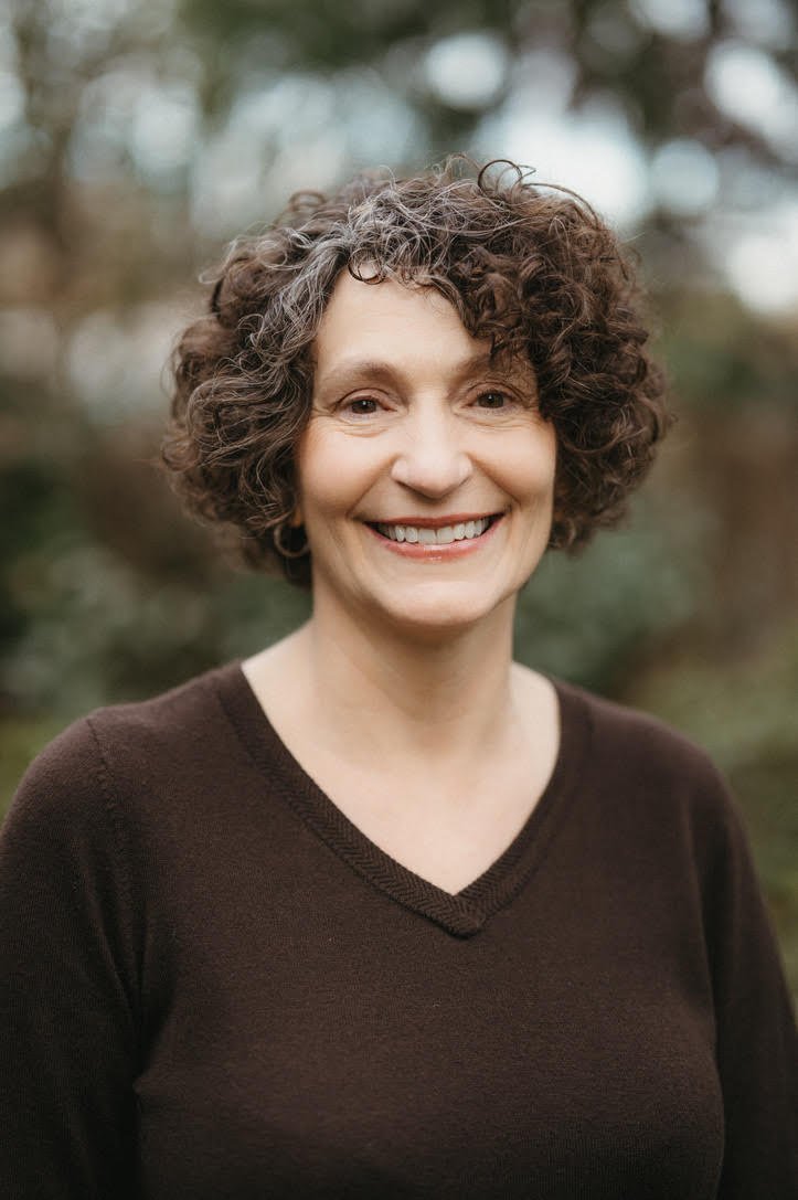 A middle-aged woman with curly brown hair smiling outdoors, wearing a dark brown V-neck sweater, blurred trees in the background.