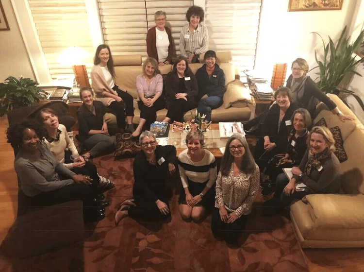 A group of fifteen women gathered in a living room, some seated on sofas and others standing behind, smiling and posing for a photo during evening book group meeting