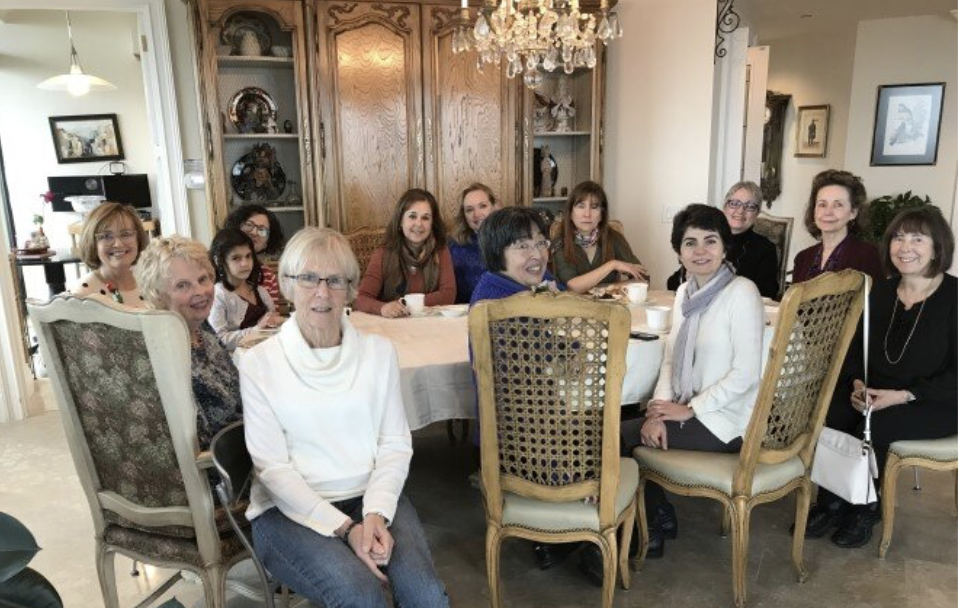 A group of women and two children gathered around a dining table in a well-lit living room with wooden cabinets and a chandelier, smiling for the photo. This is for a group that meets to speak French.