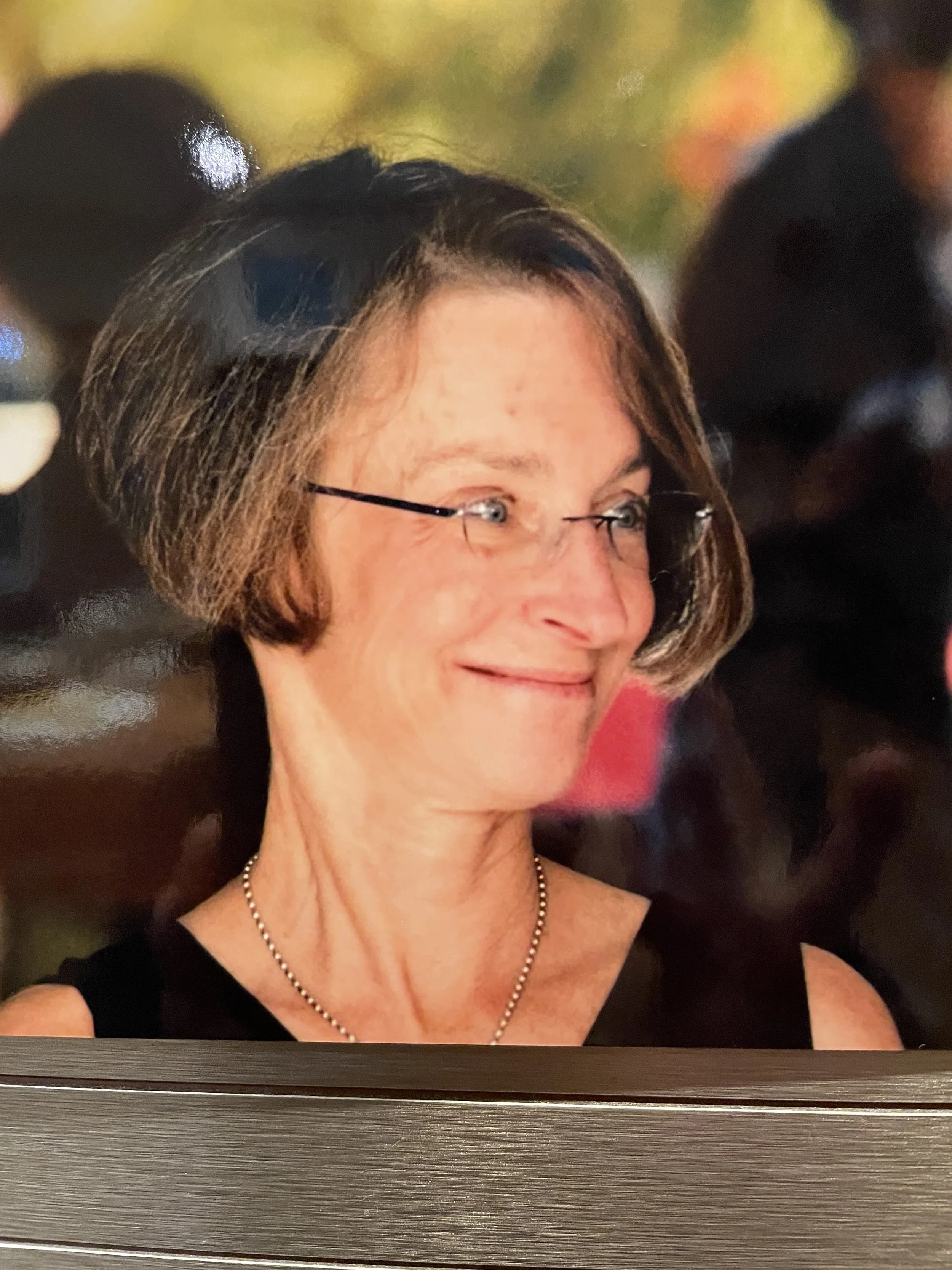 Close-up of a woman with short brown hair, glasses, and wearing a pearl necklace and black dress, smiling and looking to her right, with blurred background.