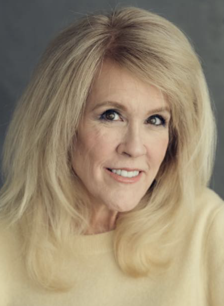 A middle-aged woman with blond hair and a tan complexion, smiling, wearing a light-colored top, against a gray background.