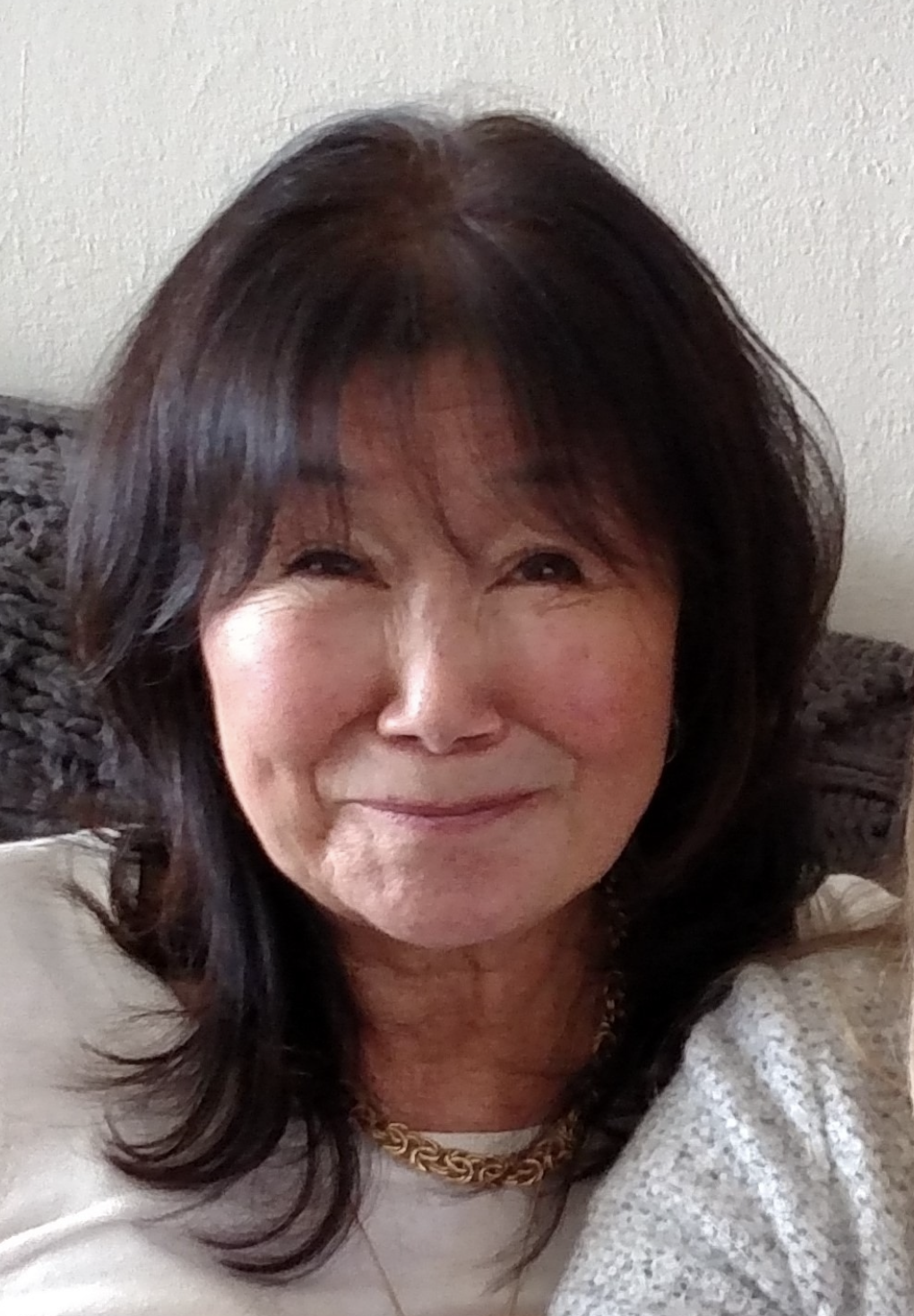 Close-up portrait of a smiling woman with dark hair, wearing a gold chain necklace, sitting on a patterned couch with a beige wall in the background.