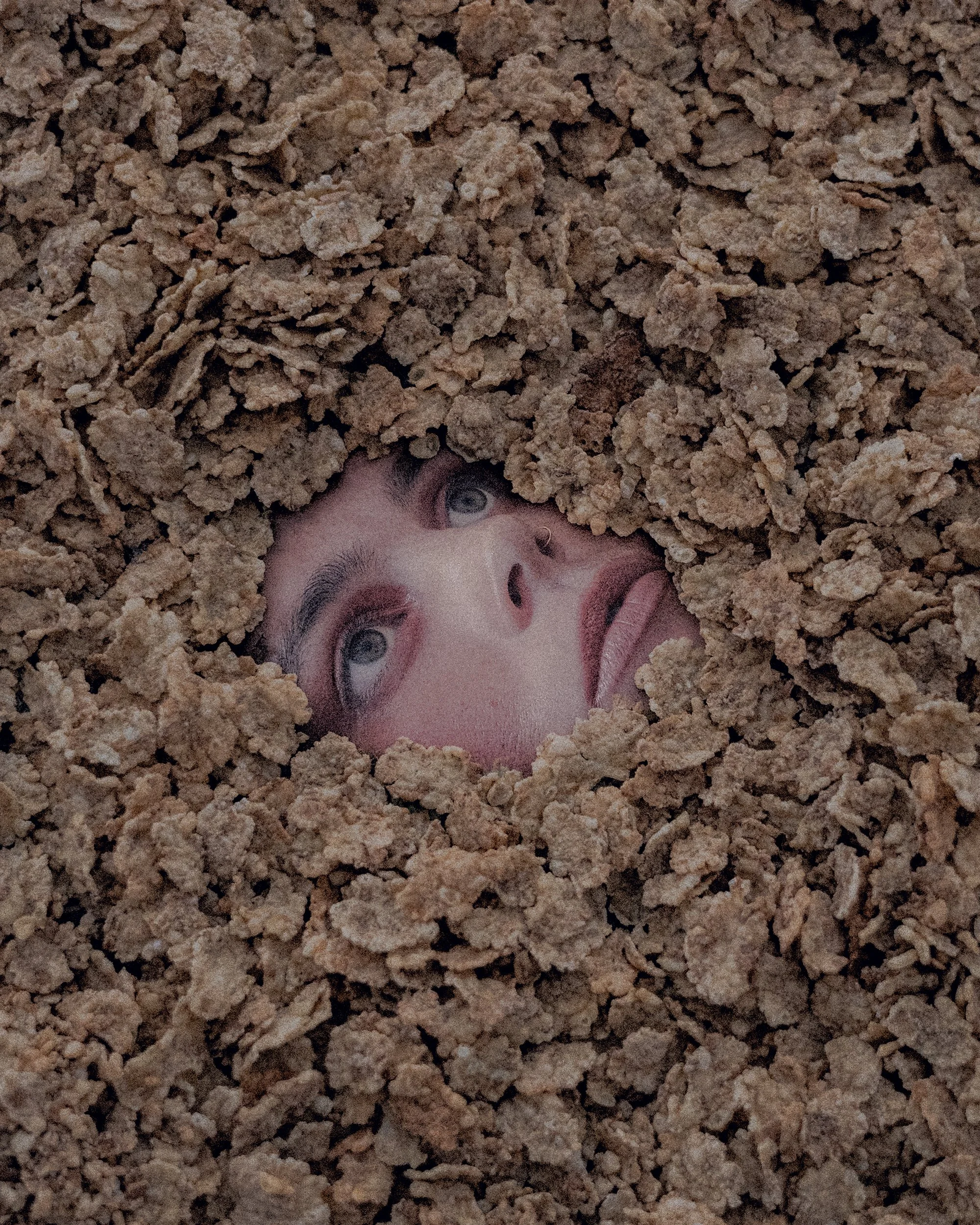 A person's face is emerging from a hole in a bed of dry, brown, cereal with only their face visible.