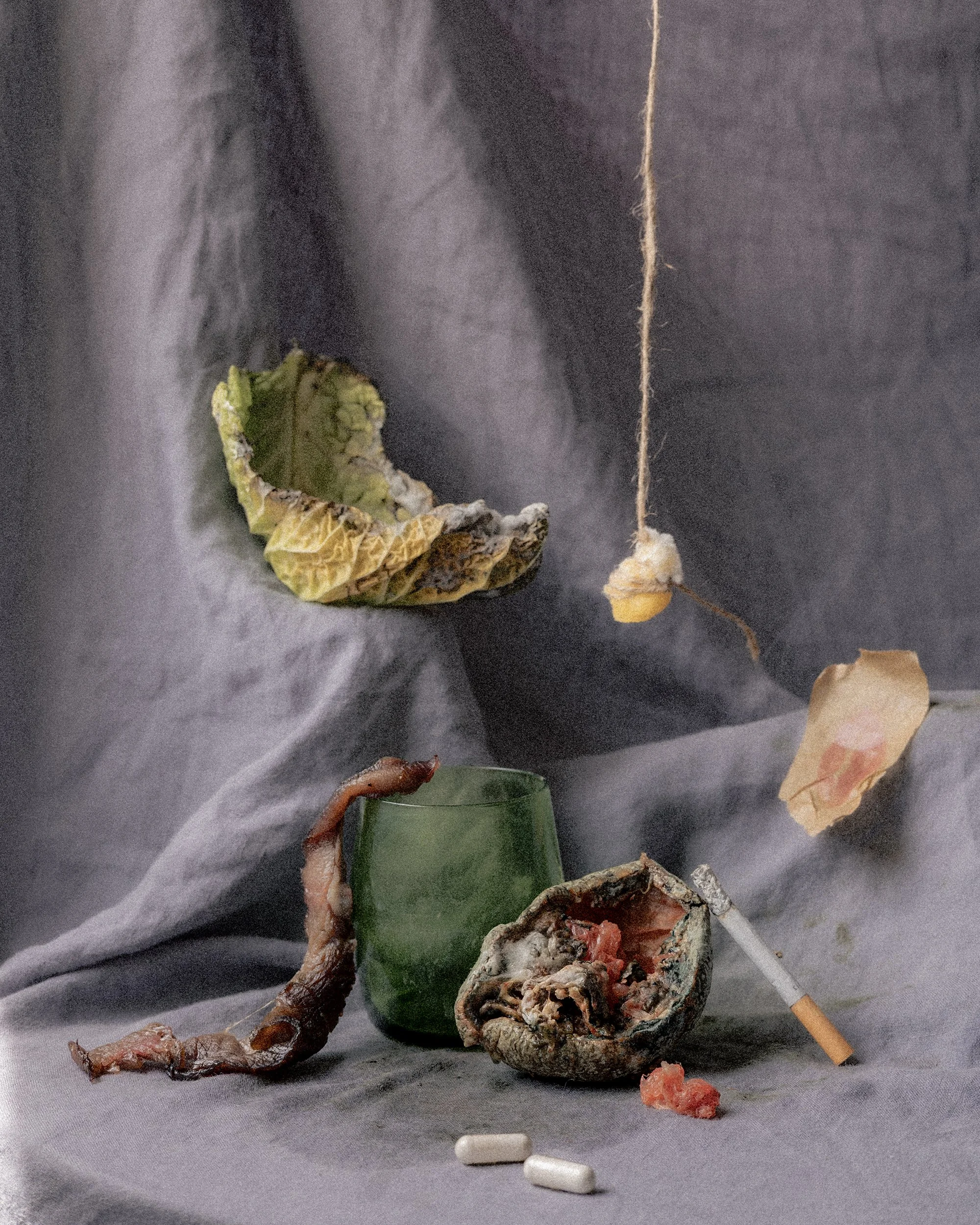 A still life arrangement of a green glass, a piece of dried meat, cigarettes, a crumpled cigarette pack, a burnt piece of fruit, a burnt leaf, and a piece of string with a small object tied at the end, all set on a gray fabric background.