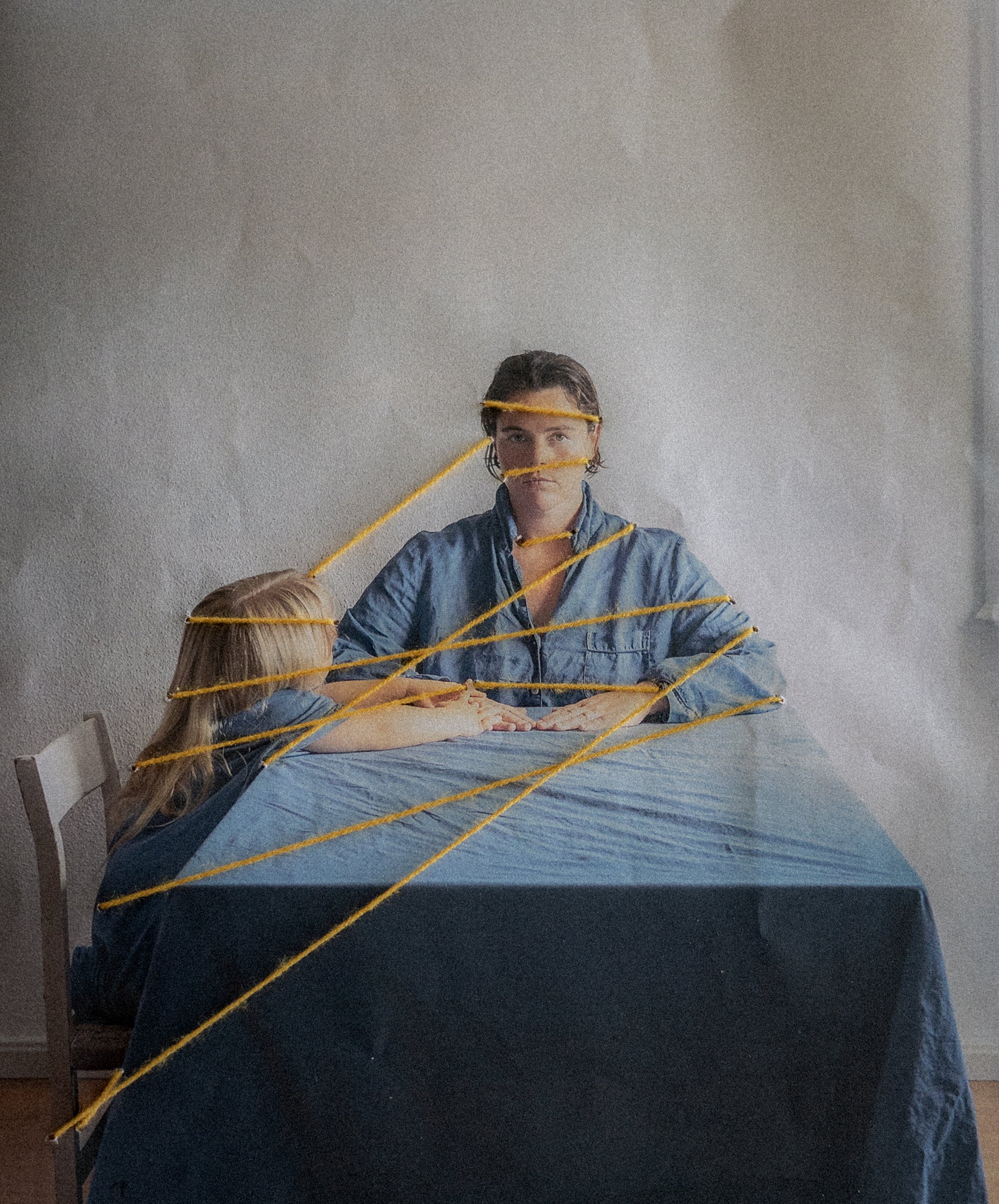 A woman and a child are sitting at a dining table covered with a blue cloth. The woman has yellow yarn wrapped around her face and arms, and the child is leaning on the table. The background is a plain, textured wall.