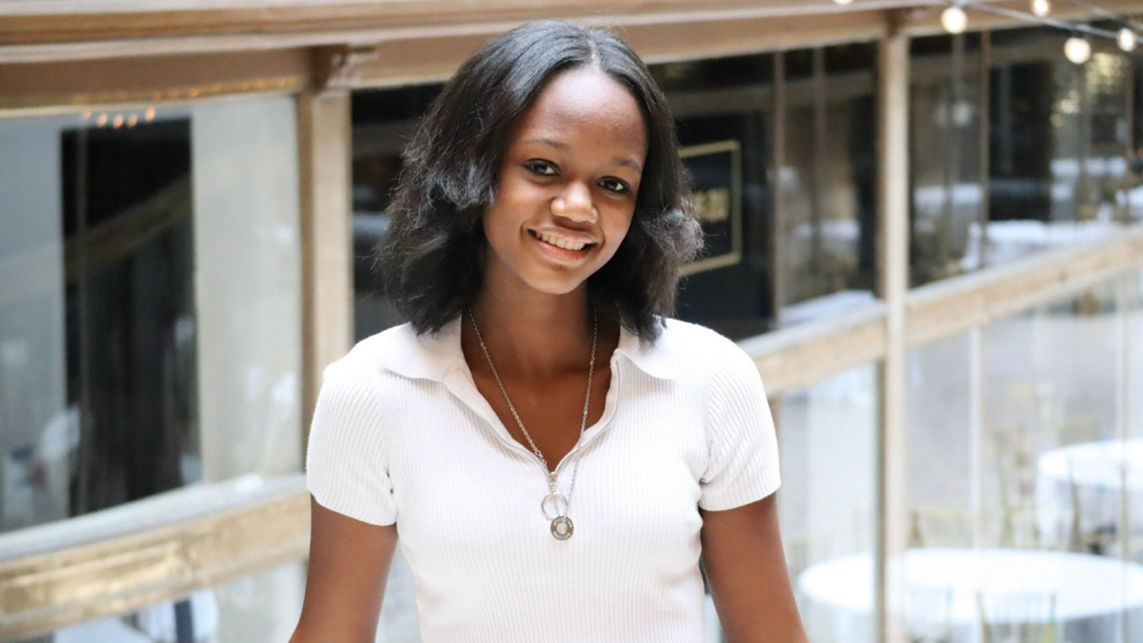 A young woman with shoulder-length black hair, smiling, wearing a white short-sleeved shirt and a necklace, standing indoors with modern building interior in the background.