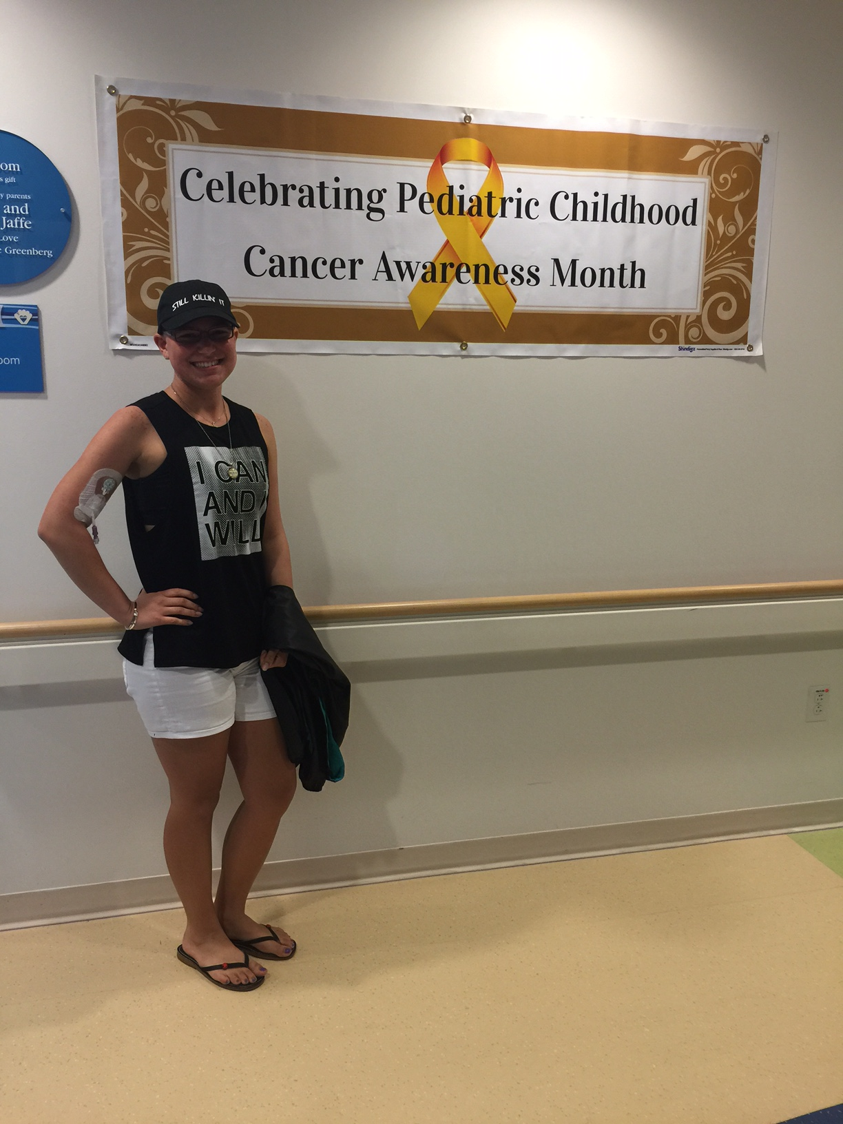 A woman standing in front of a banner that reads 'Celebrating Pediatric Childhood Cancer Awareness Month' with a yellow ribbon symbol. She is smiling, wearing a black cap, black shirt, white shorts, and flip-flops, with an IV patch on her arm and holding a jacket.