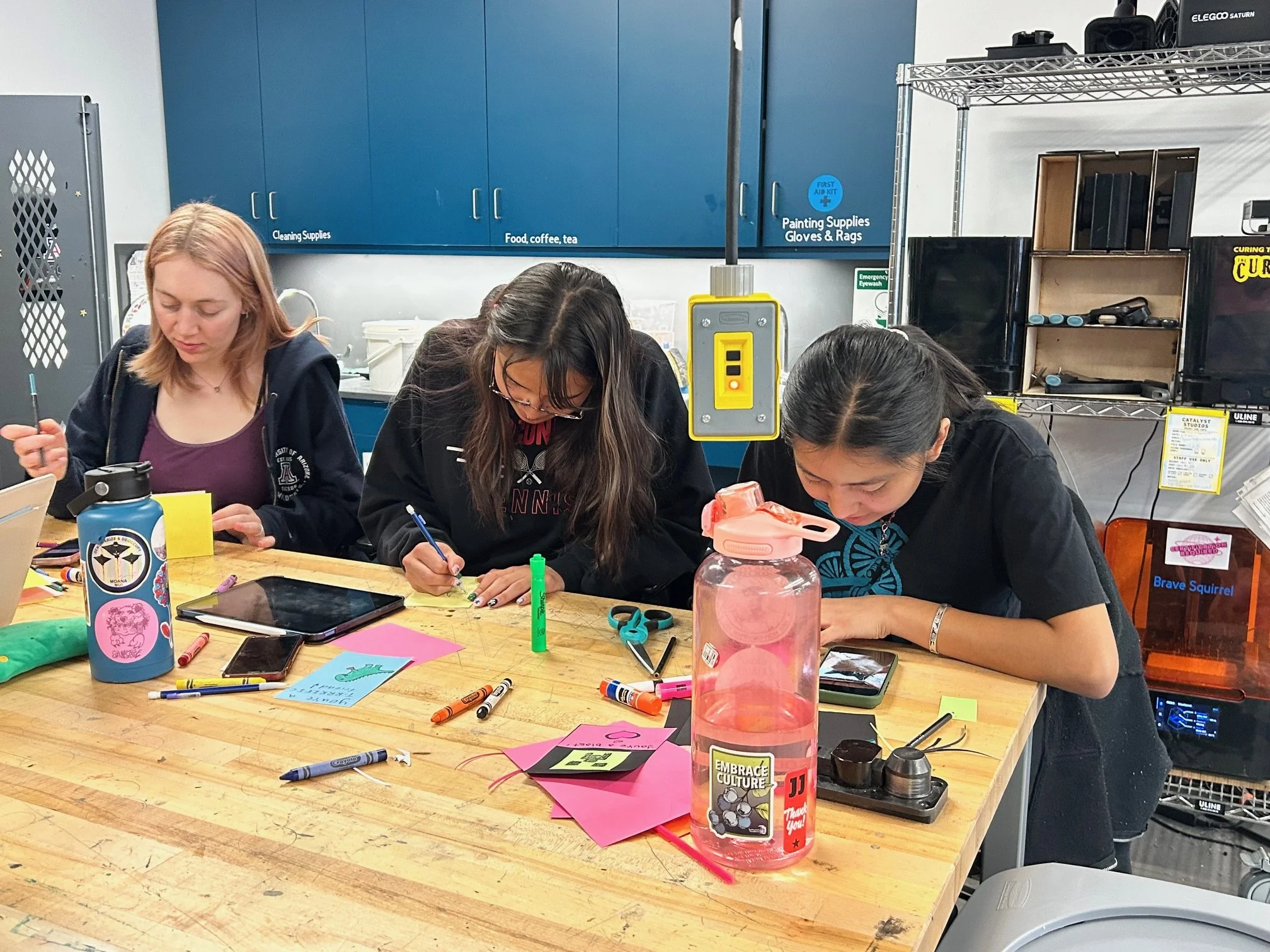 Three women are sitting at a cluttered table engaged in arts and crafts activities. The table has markers, papers, and a tablet. Behind them is a blue cabinet with labeled sections for supplies, and a wire rack with various items including speakers and books. One woman is writing, another is working with paper, and the third is focused on her work.