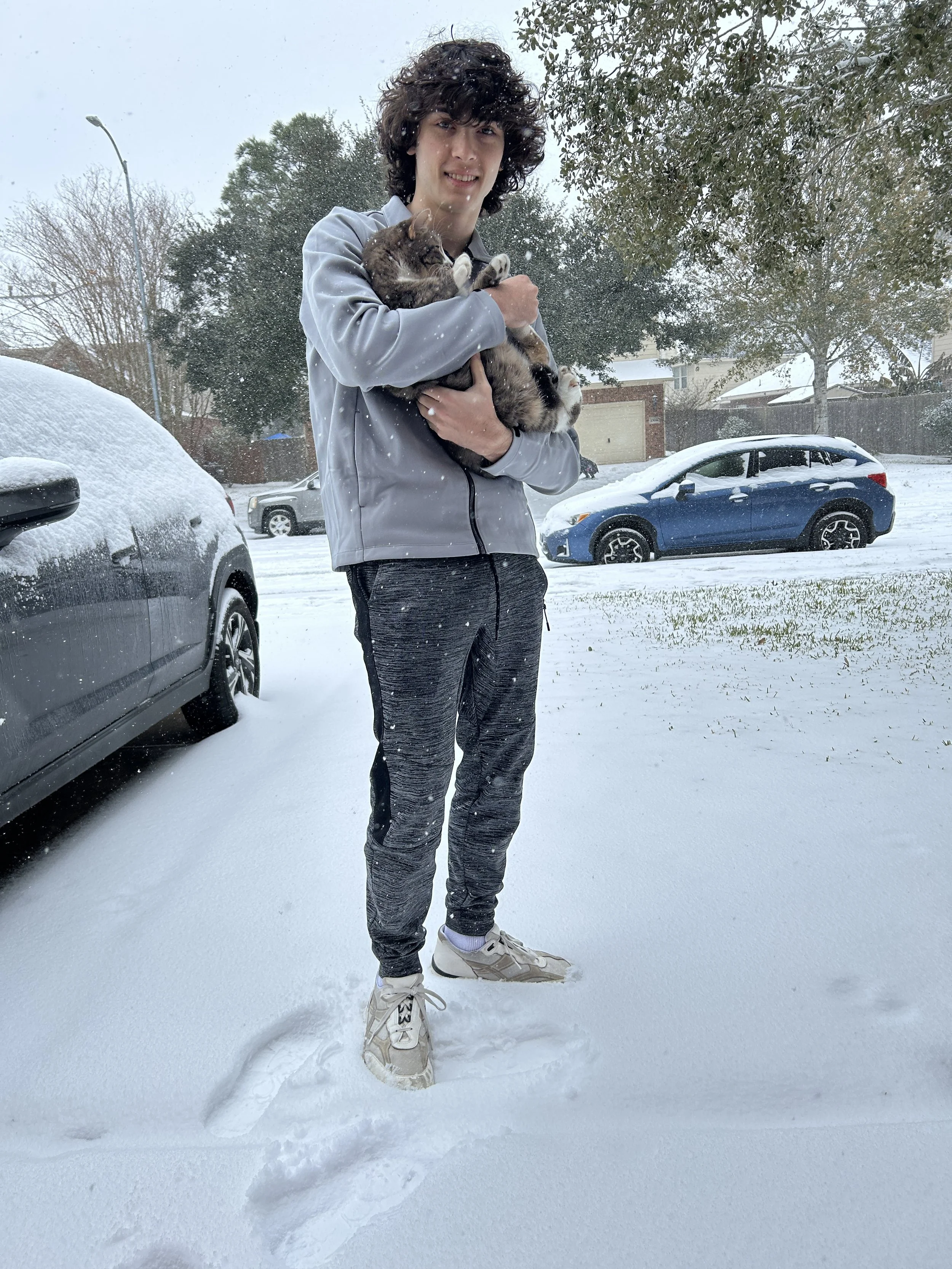 A young man with curly hair wearing a gray jacket, black pants, and white sneakers holds a tabby cat in a snowy outdoor scene with parked cars and trees in the background.