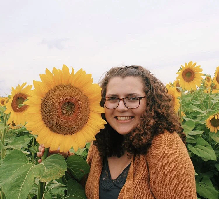 A woman with curly hair and glasses smiling while standing among sunflowers in a sunflower field.