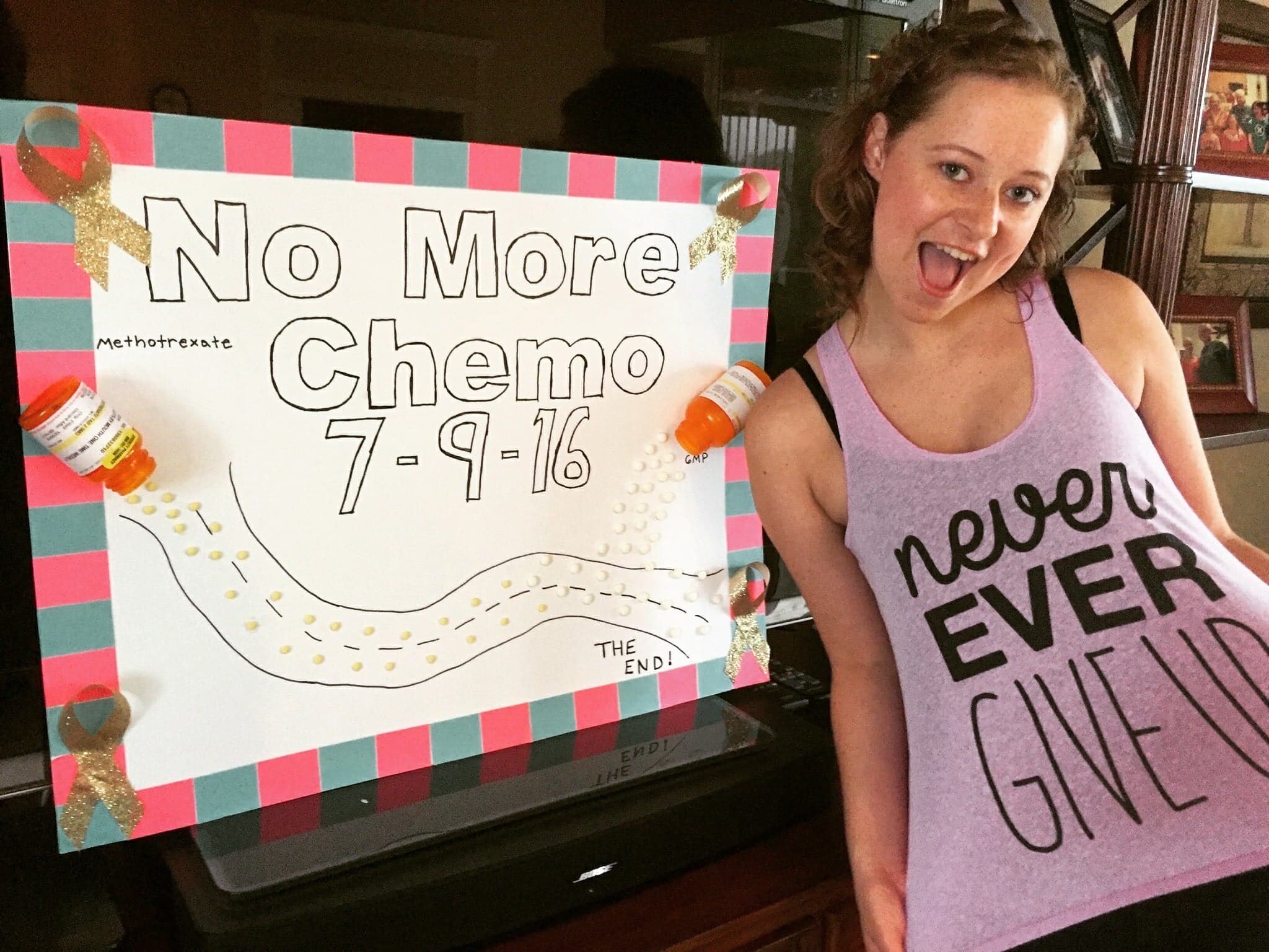 A woman with curly hair holding a sign that reads 'NO More Chemo 7-9-16'. The sign decorated with gold ribbons and pills, with a drawing of a bottle pouring out pills labeled 'Methotrexate' and the words 'THE END!'