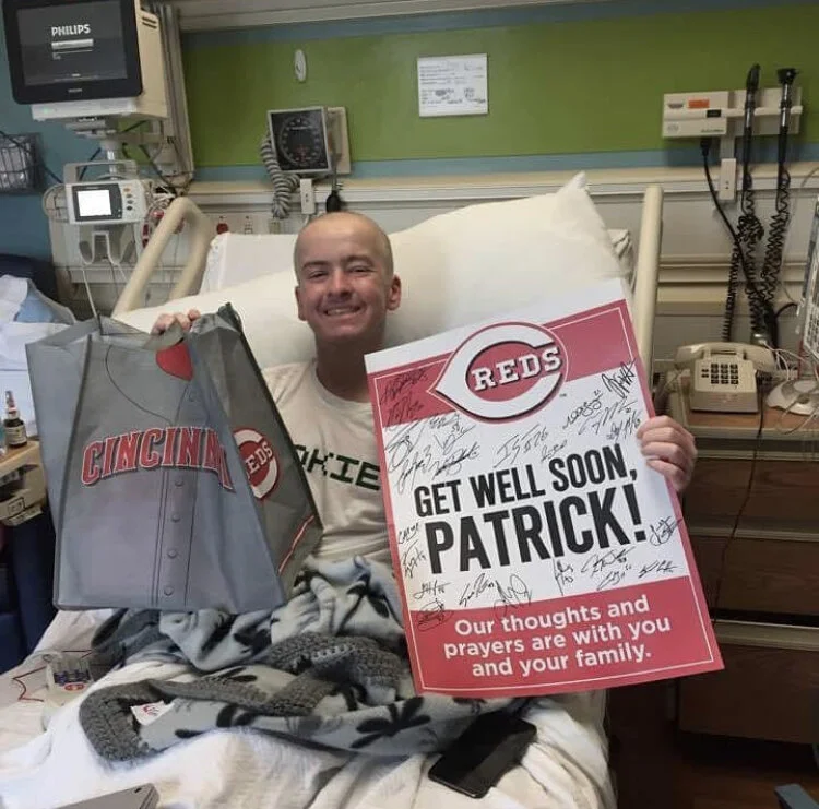 A man in a hospital bed holding a Cincinnati Reds gift bag and a sign that says, "Get Well Soon Patrick!" with messages of support and hearts.