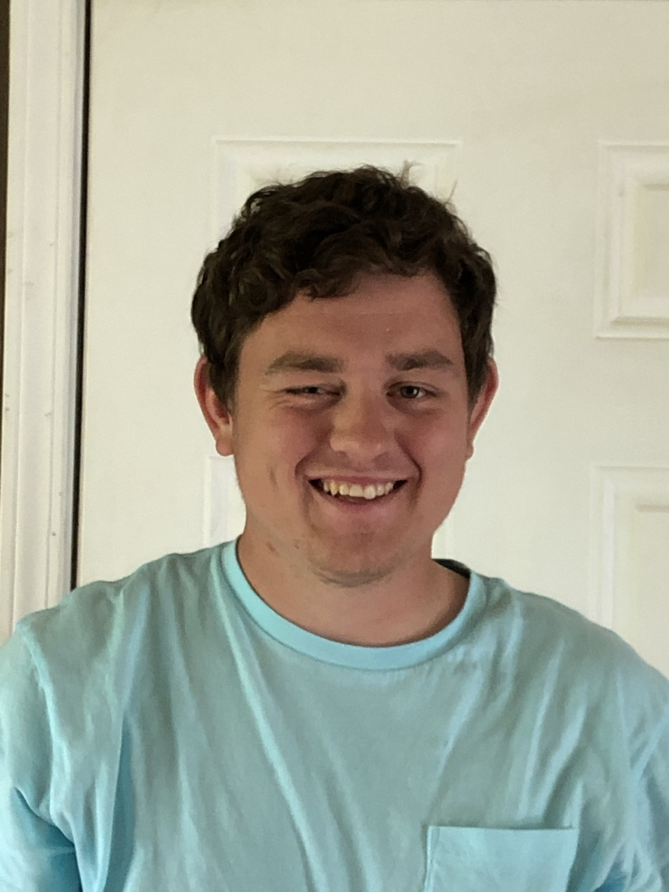 A young man smiling and winking, wearing a light blue T-shirt, standing in front of a white door.