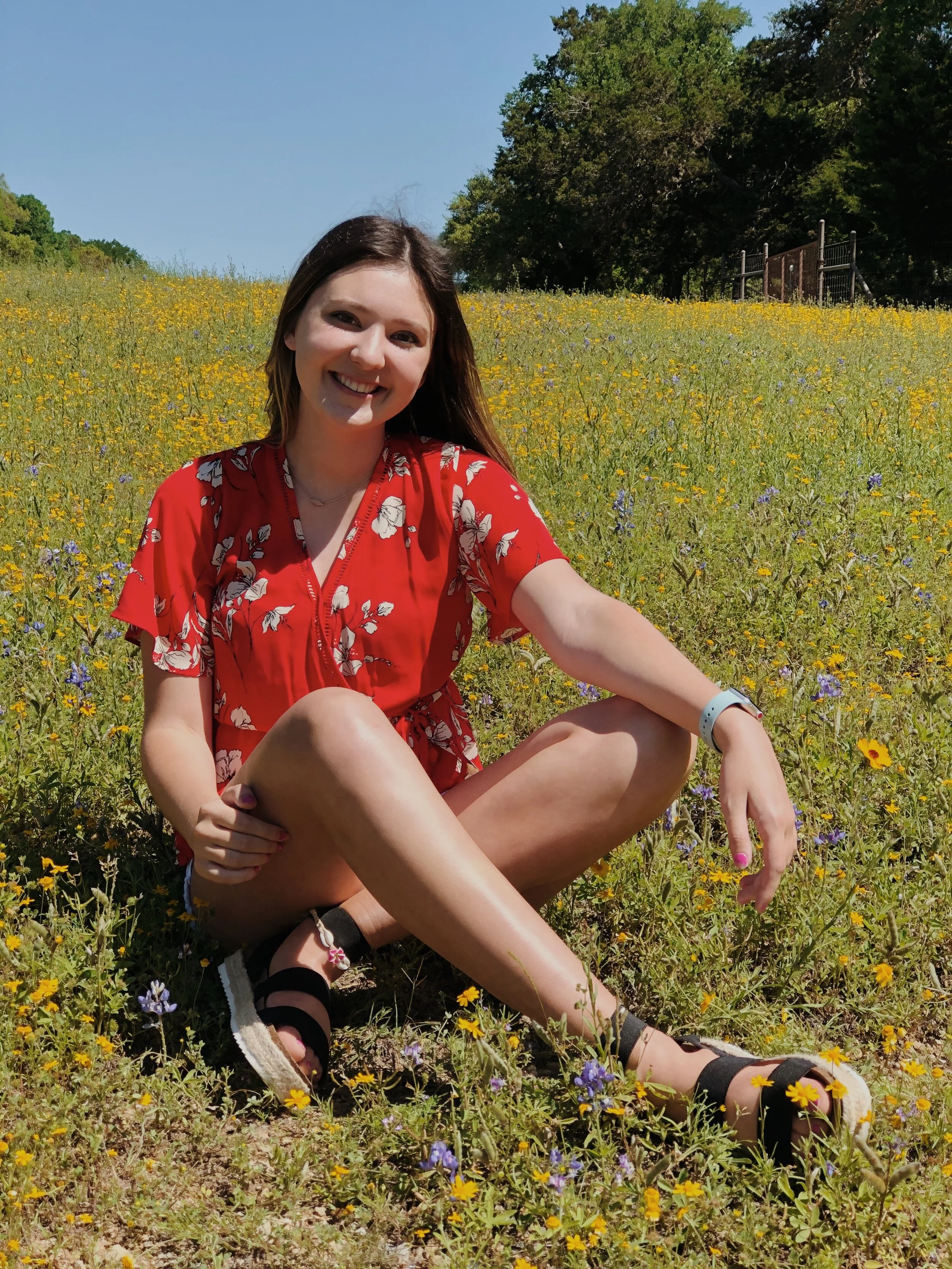 A young woman with long brown hair, wearing a red floral dress and sandals, sitting in a field of yellow and purple wildflowers on a sunny day with a blue sky and green trees in the background.