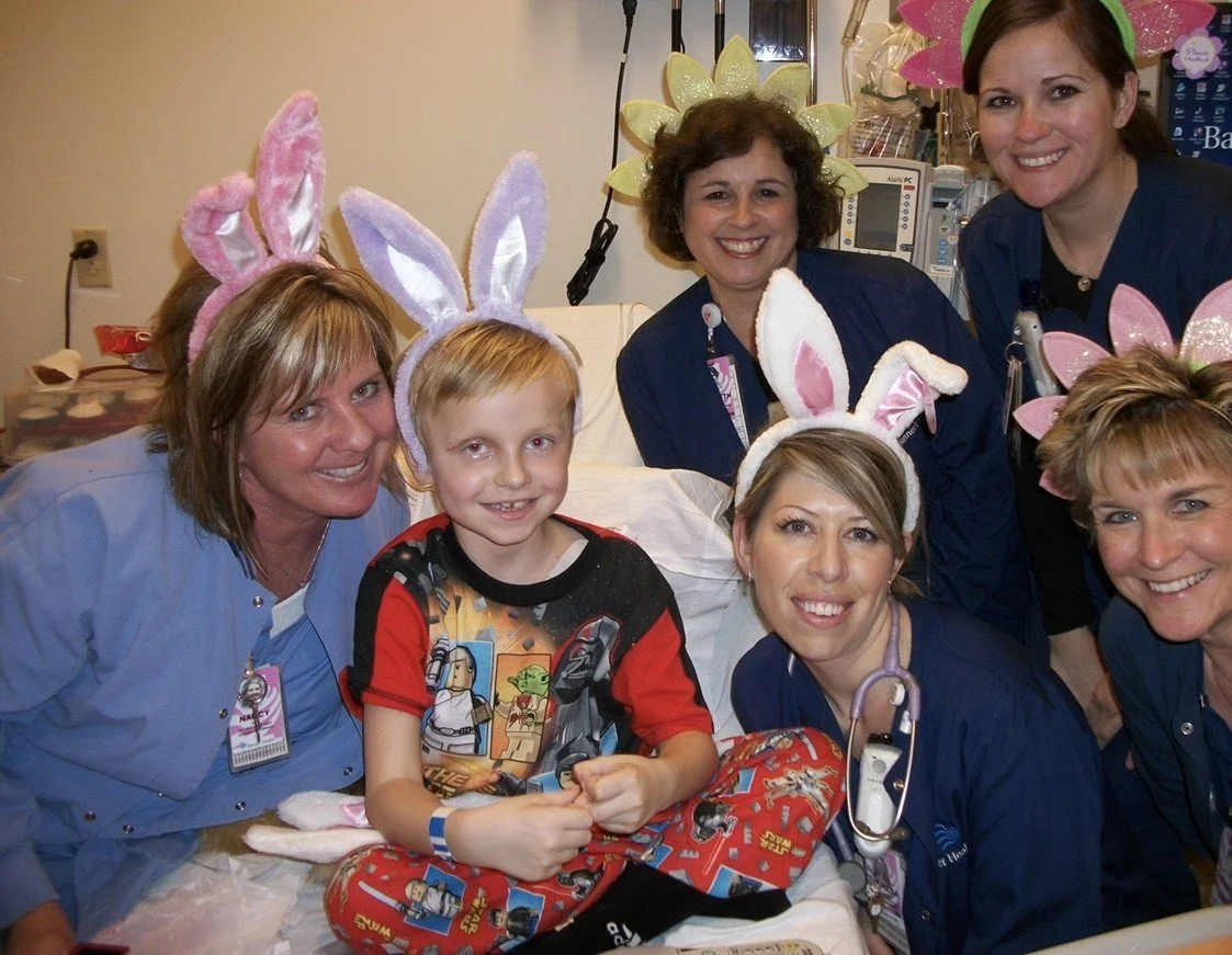 A young boy wearing arm and leg braces, sitting in a hospital bed, surrounded by five women and a girl, all wearing bunny ears and pink or yellow flowers headbands, smiling during a festive event.