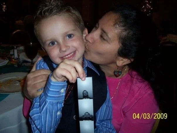 A woman is kissing a young boy on the cheek at a dinner table, both smiling. The boy is wearing a blue shirt with a black vest and a black and white tie. The woman is wearing a pink top and heart-shaped earrings. There is a timestamp in the lower right corner reading 04/03/2009.
