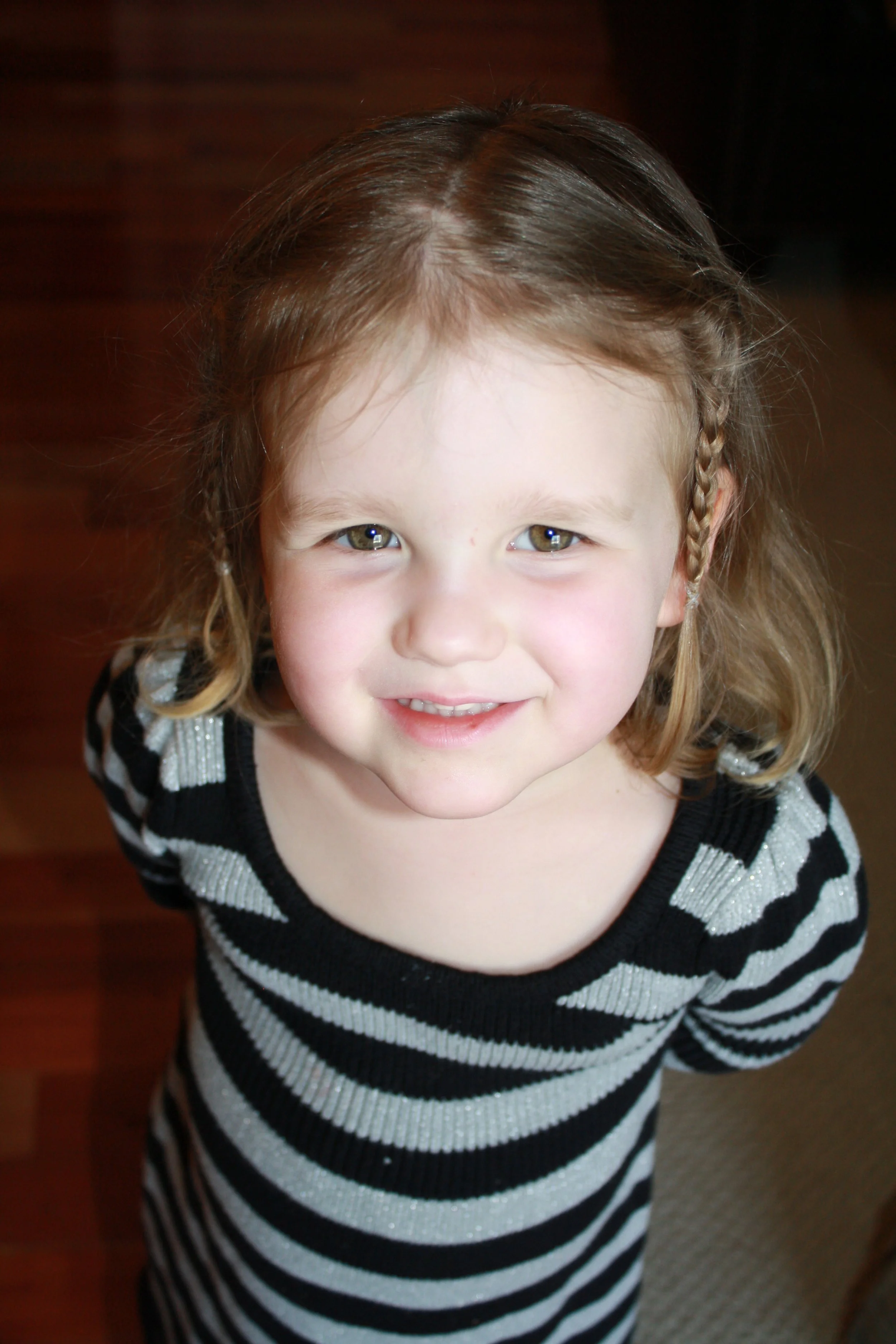 A young girl with light-colored hair styled in braids, wearing a black and white striped sweater, smiling upward.