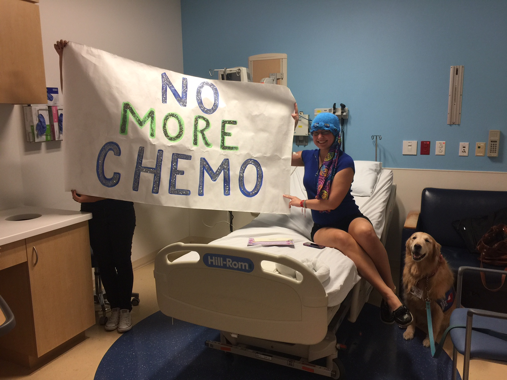 A woman in a hospital bed wearing a blue hat, smiling, pointing at a large white poster that reads 'NO MORE CHEMO' in glittery letters. Another person holds the poster, and a golden retriever dog is sitting beside the bed. The hospital room has medical equipment and a blue and white color scheme.