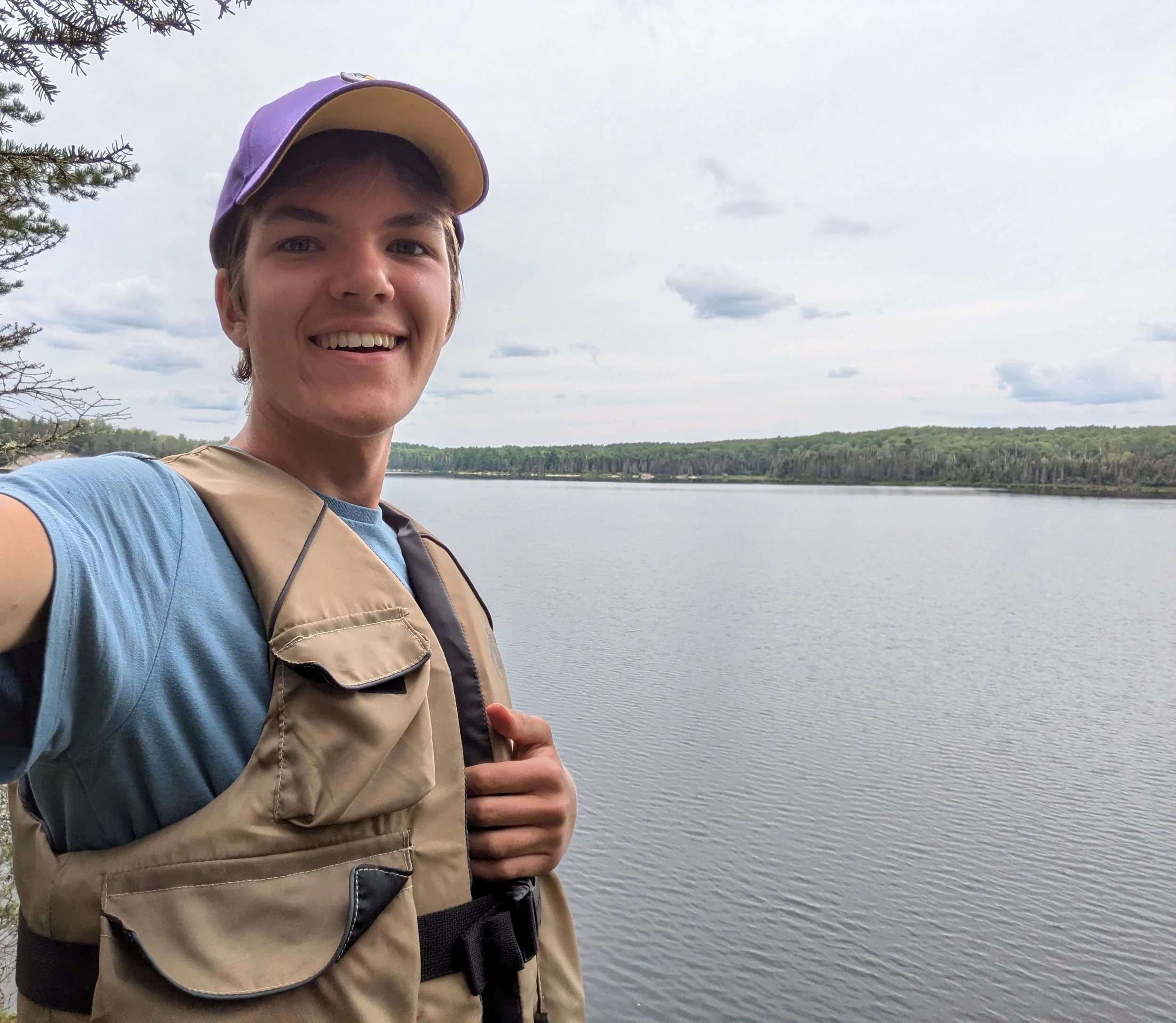 A young man taking a selfie outdoors near a river with a forested landscape in the background, wearing a purple and yellow cap, a beige vest, and a blue shirt.