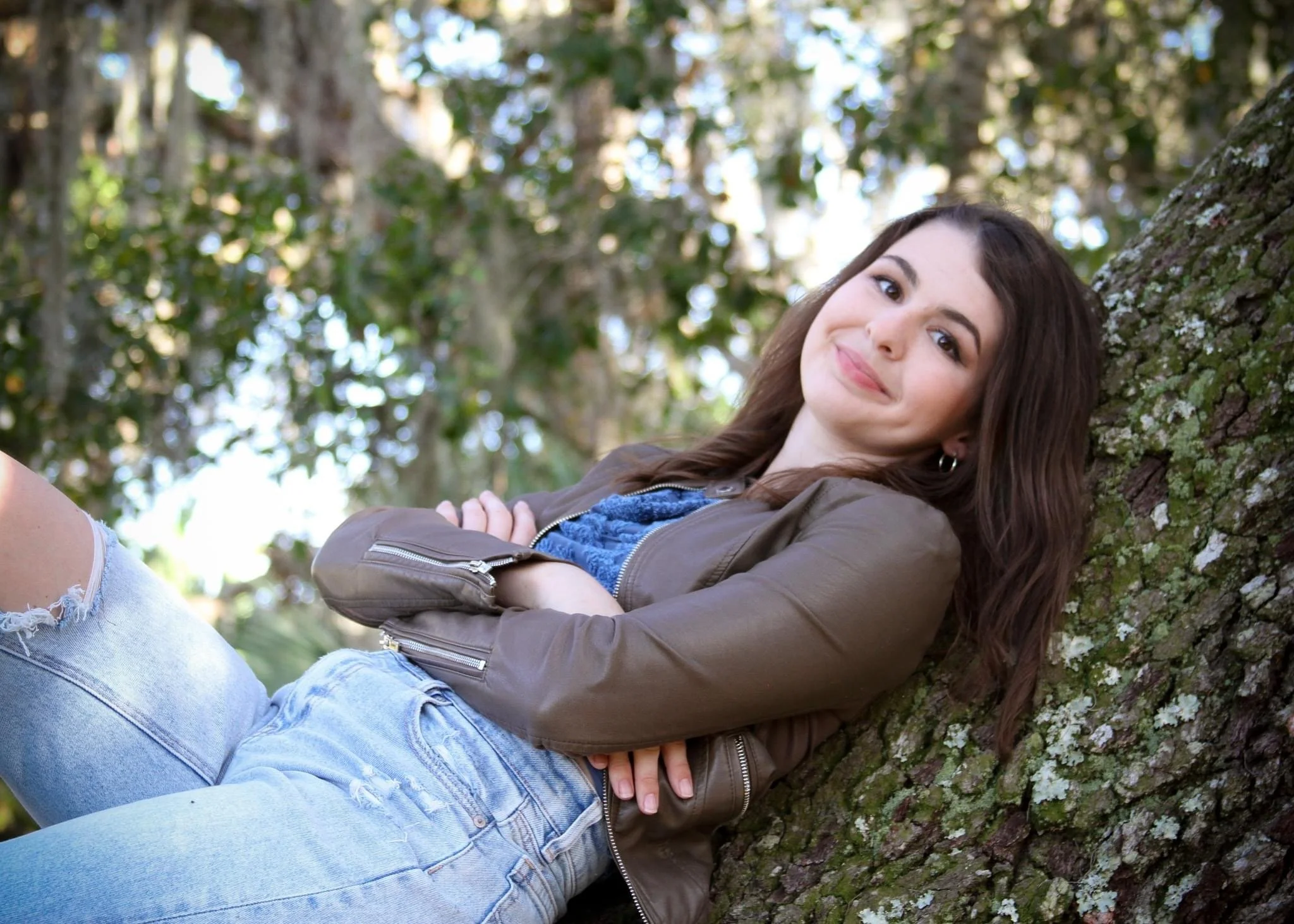 A woman with long brown hair is lying on a tree trunk outdoors, smiling at the camera. She is wearing a brown jacket and ripped light blue jeans, and there are green leaves and branches in the background.
