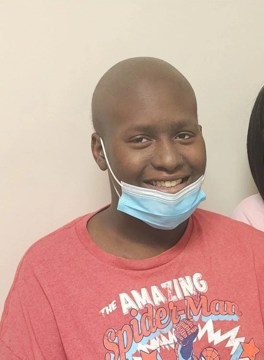 A young boy with a shaved head smiling, wearing a blue face mask and a red Spider-Man t-shirt.