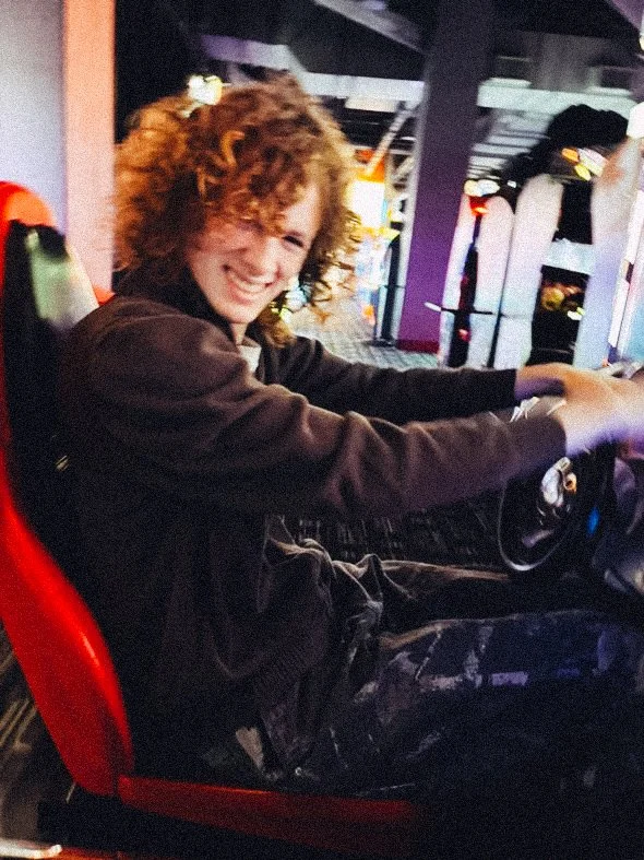 A smiling person with curly hair sitting in a racing ride with a red seat, holding a steering wheel, in an arcade or amusement park.