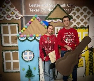 Two young people standing in front of a festive backdrop with a house made of craft materials. One is holding a guitar prop. They are smiling and dressed casually.